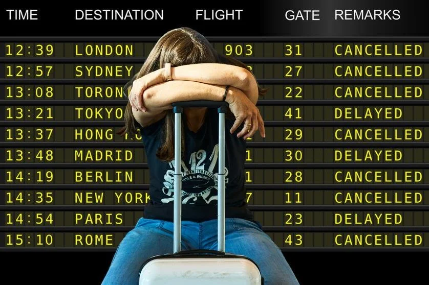 Frustrated traveler sitting with luggage in front of an airport departures board showing delayed and canceled flights.