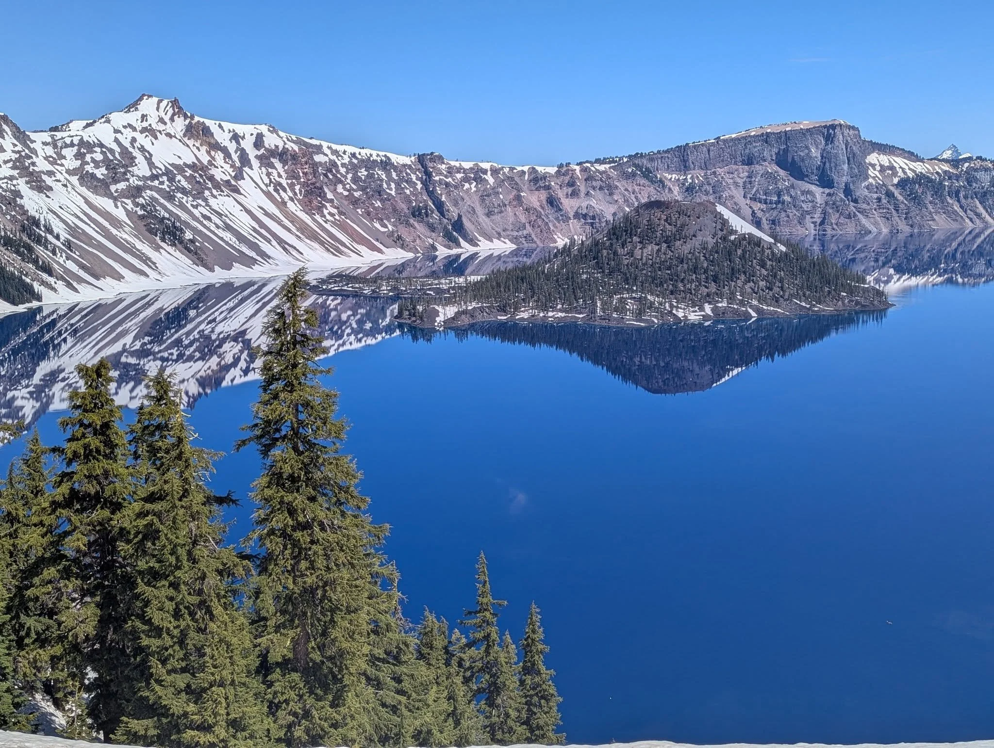 Crater Lake in Oregon on a clear late-May day, with snow-covered volcanic cliffs and Wizard Island reflected in the deep blue water.