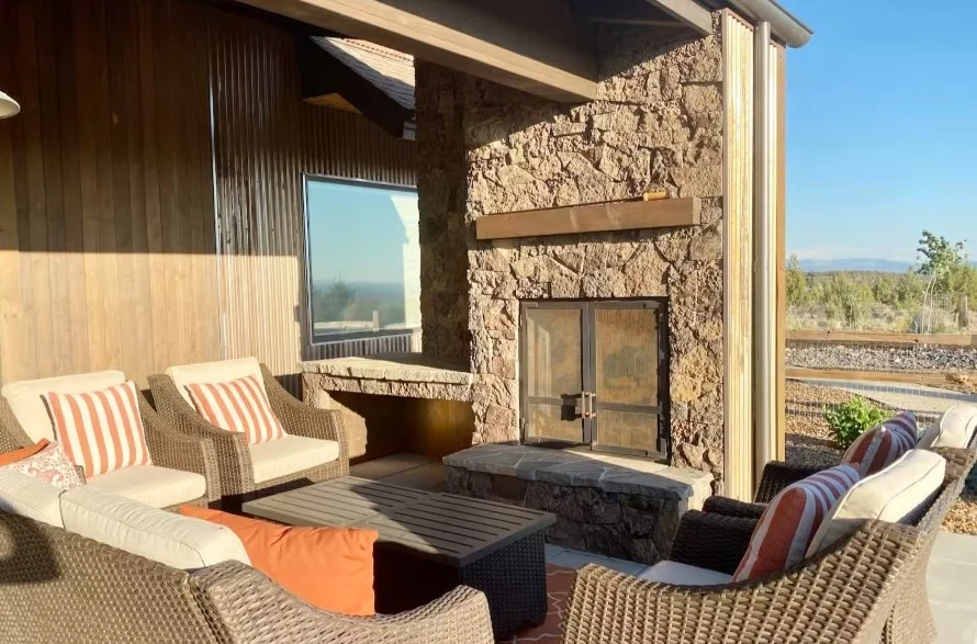 Comfortable wicker seating arranged around a low table beside a stone outdoor fireplace at Panoramic Mountain View Oasis in Powell Butte.