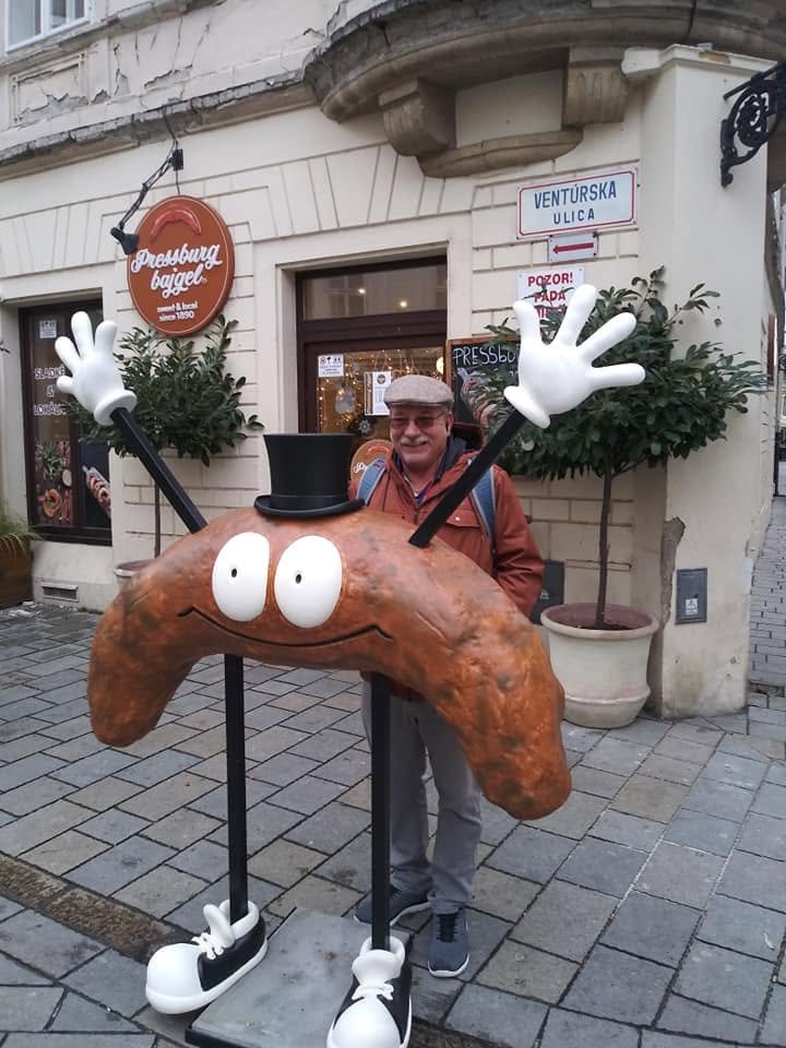 Traveler posing beside a whimsical giant croissant character outside a bakery on a pedestrian street in Bratislava, Slovakia.