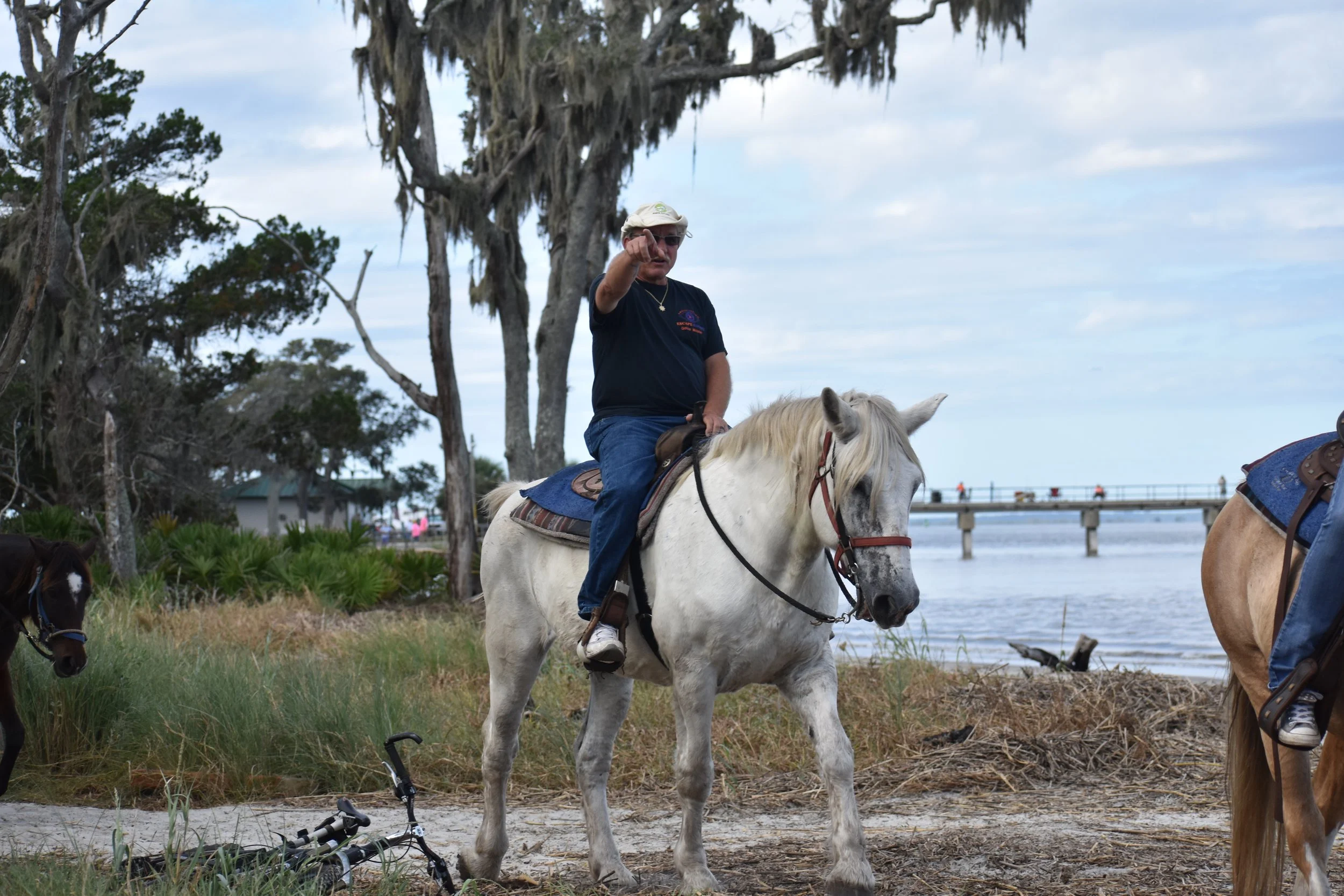 Brian riding a white horse along the Jekyll Island shoreline, with Spanish moss hanging from the trees and a fishing pier extending over the water in the background.