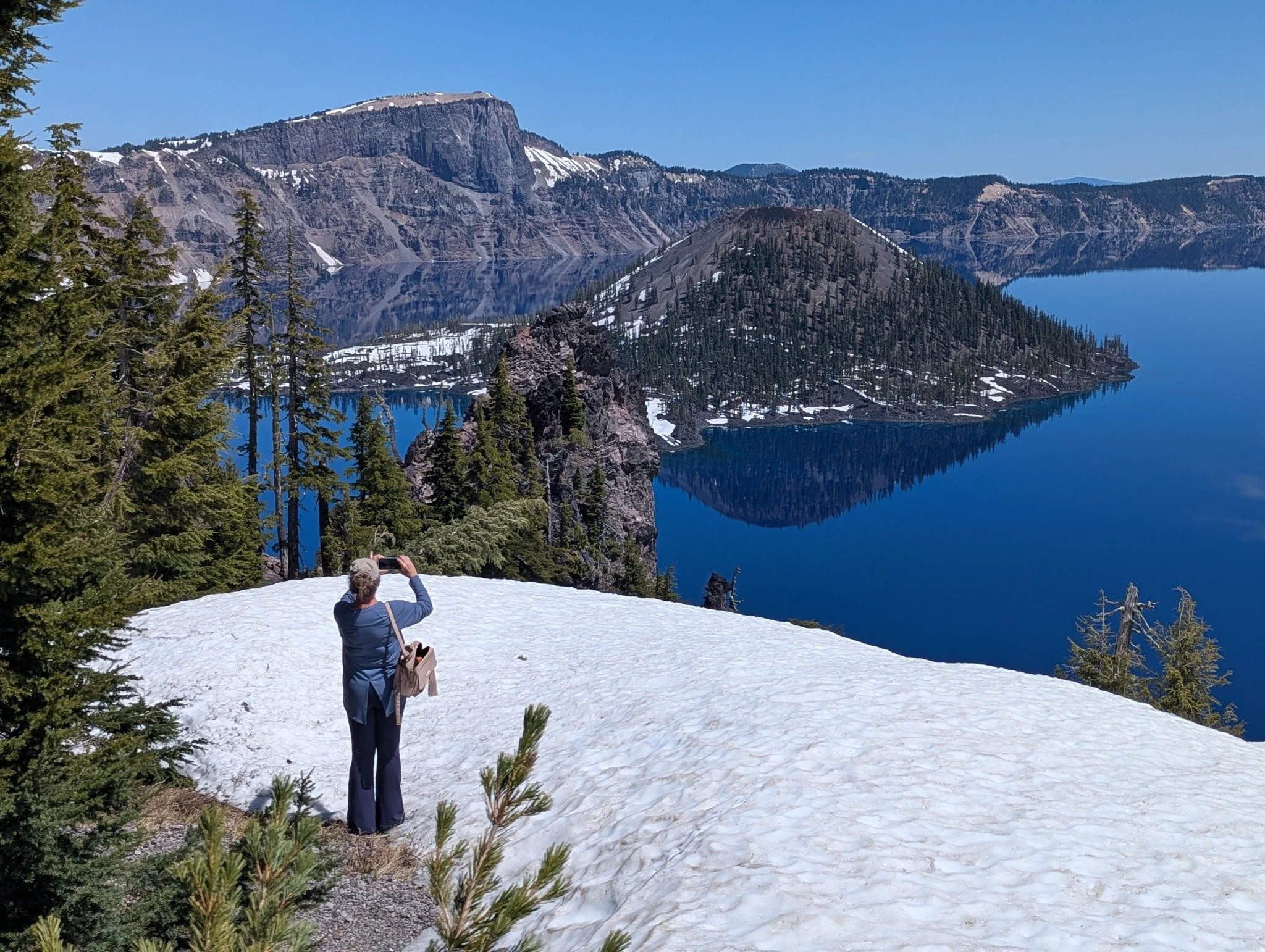 Gina photographing Wizard Island and its reflection in the still blue waters of Crater Lake, with snow covering the overlook and volcanic cliffs surrounding the lake.