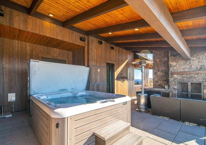 Hot tub beneath a covered timber patio at a Powell Butte vacation rental, with wooden beams, outdoor seating, and a stone fireplace.