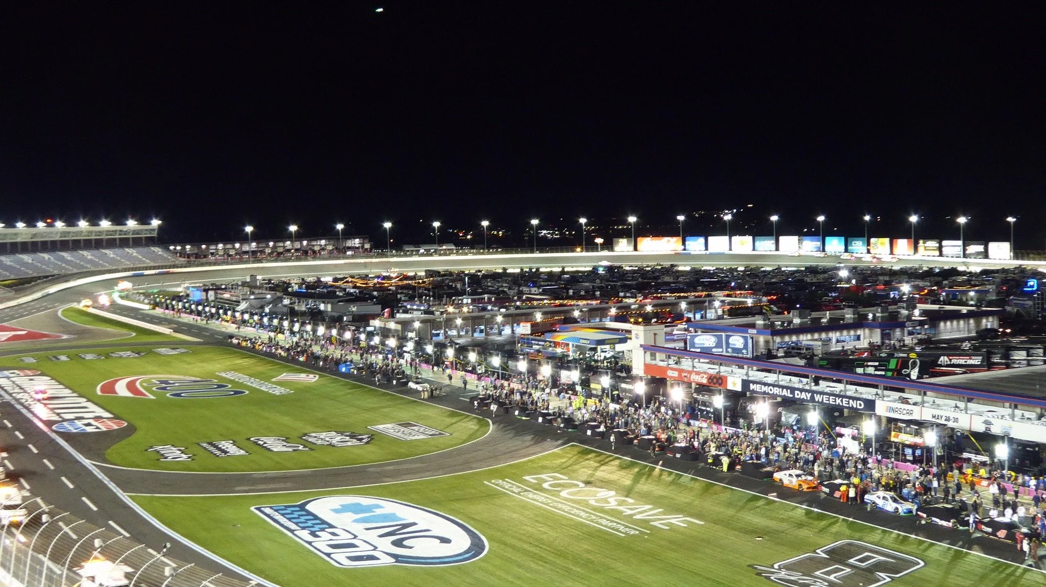 Nighttime Panasonic Lumix ZS50 photograph of Charlotte Motor Speedway showing illuminated pit road, the racing surface, and surrounding grandstands.