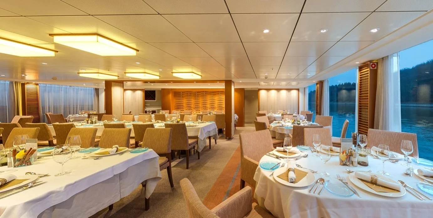 Viking Ingvi dining room set for a meal, with white tablecloths, elegant place settings, comfortable chairs, and panoramic river views.