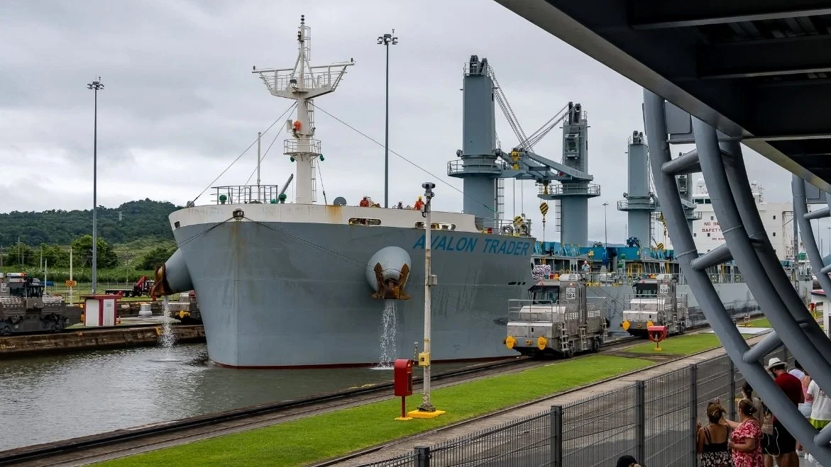 Cargo ship Avalon Trader passing through the Panama Canal's Miraflores Locks, with electric towing locomotives and lock infrastructure alongside the vessel.