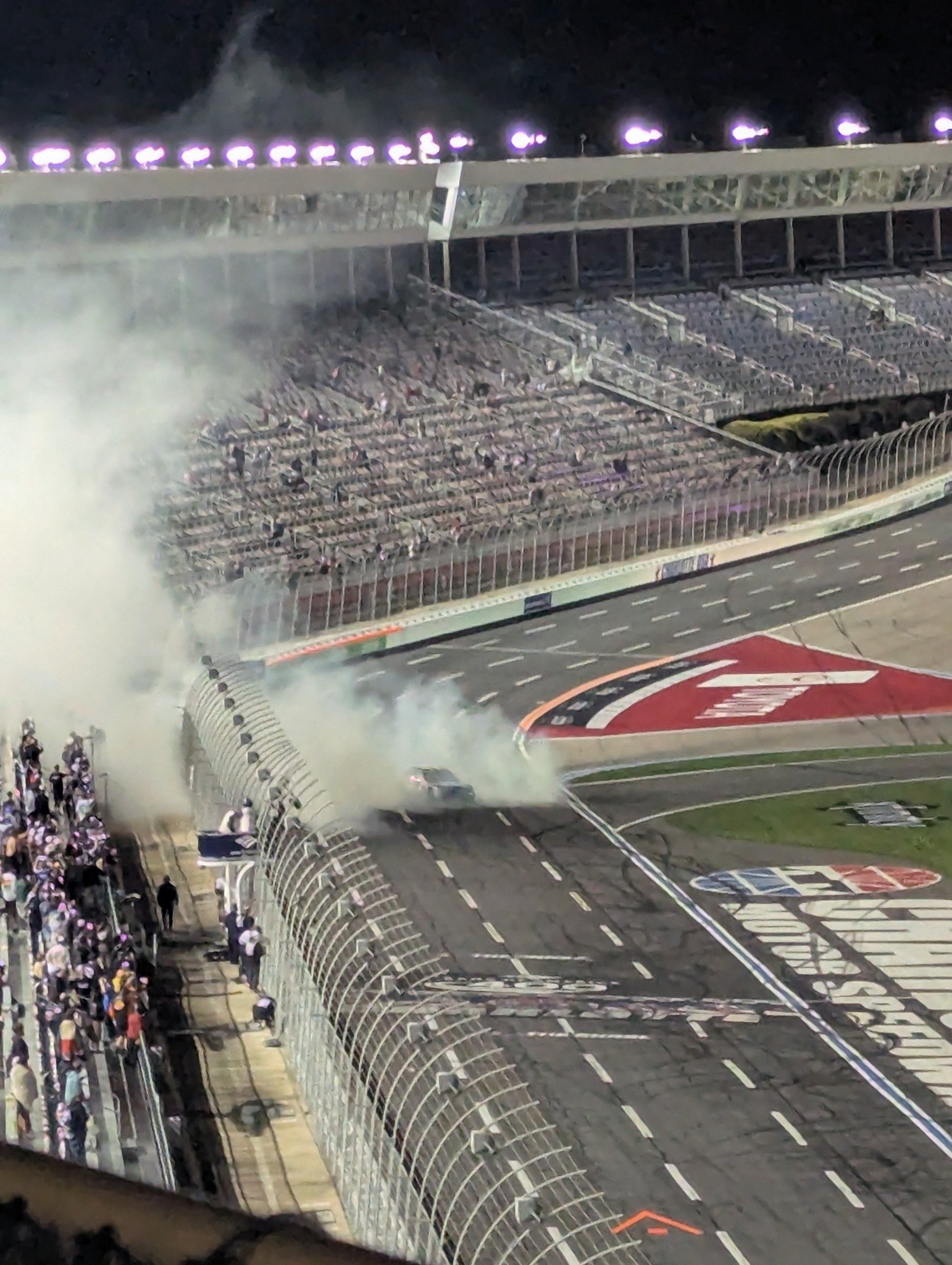 Google Pixel 7 photograph of a nighttime NASCAR burnout at Charlotte Motor Speedway, with a race car partly obscured by tire smoke beneath the track lights.
