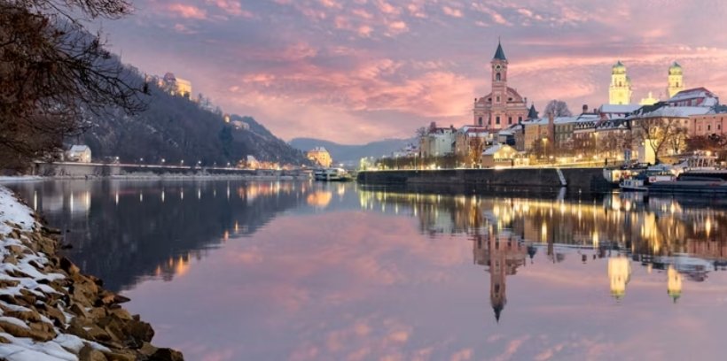 Winter evening on the Danube in Passau, Germany, with snow along the riverbank, bare trees, historic waterfront buildings, and warm lights reflected in the water.