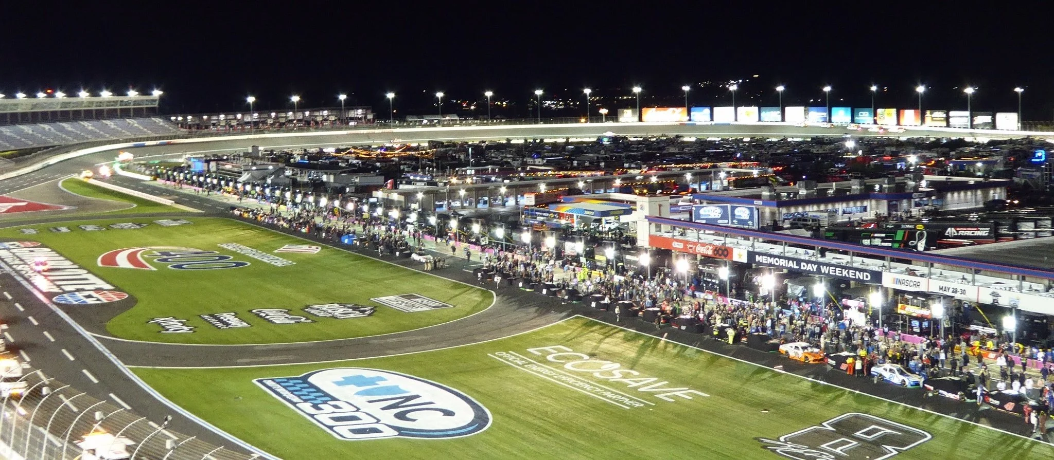 Nighttime view of Charlotte Motor Speedway during a NASCAR race weekend, featuring the brightly illuminated pit road, infield garages, and banked racetrack.