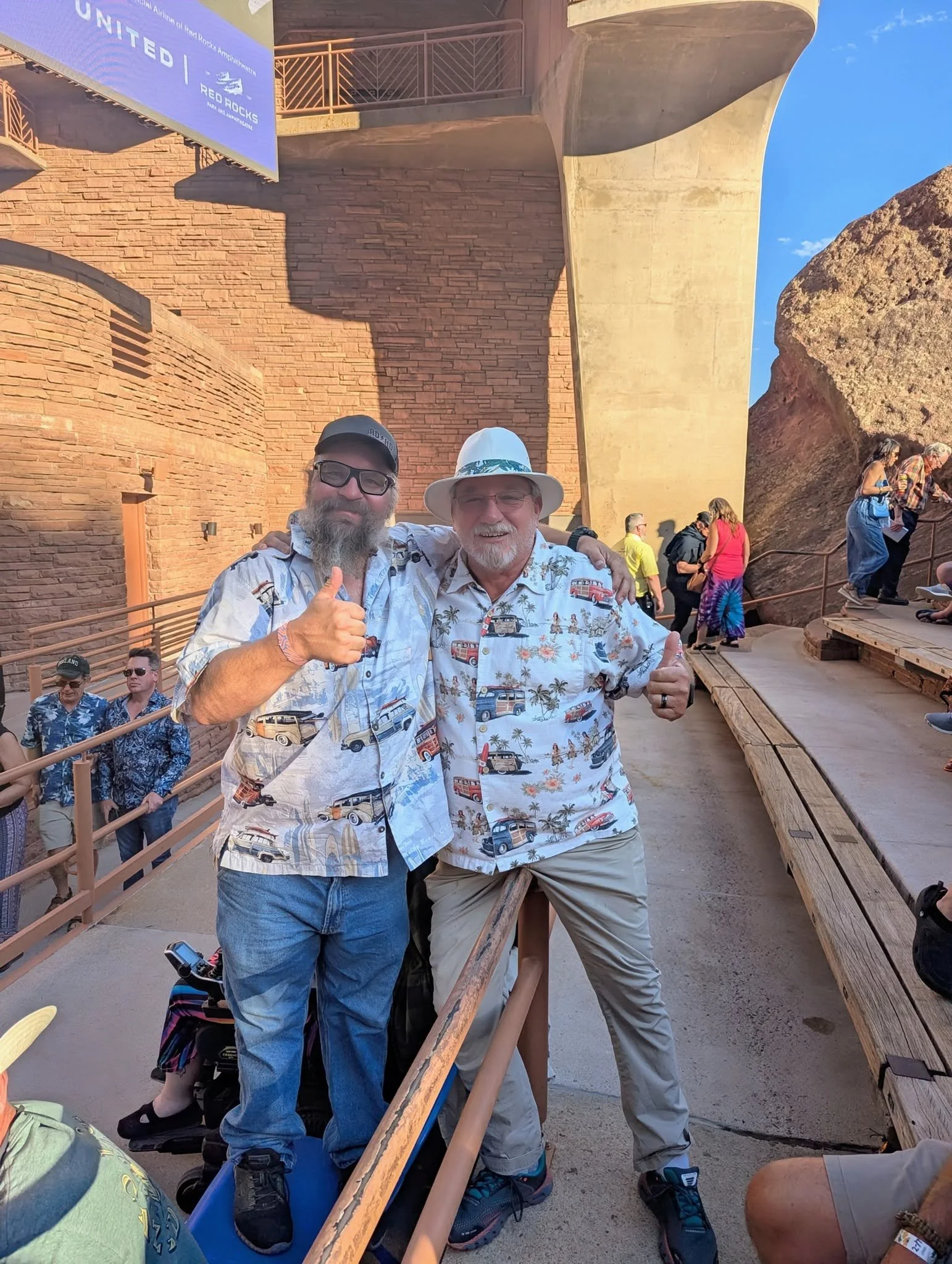 Two concertgoers wearing matching tropical shirts give thumbs-up at a Beach Boys concert at Red Rocks Amphitheatre in Colorado.