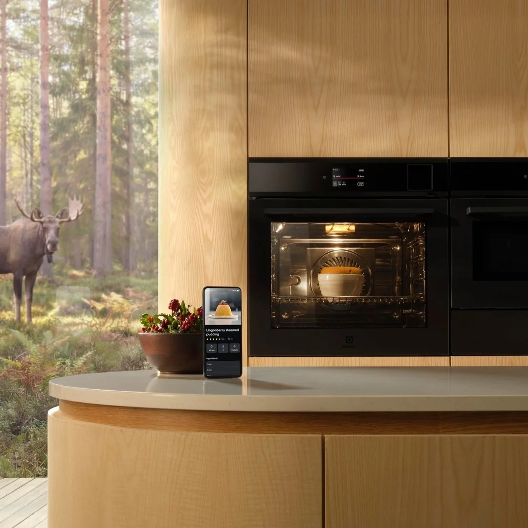 A modern kitchen with wooden cabinets and a built-in black oven. On the counter, there is a bowl of berries and a smartphone displaying a recipe for lingonberry steamed pudding. Outside the window, a moose is visible in a forest.