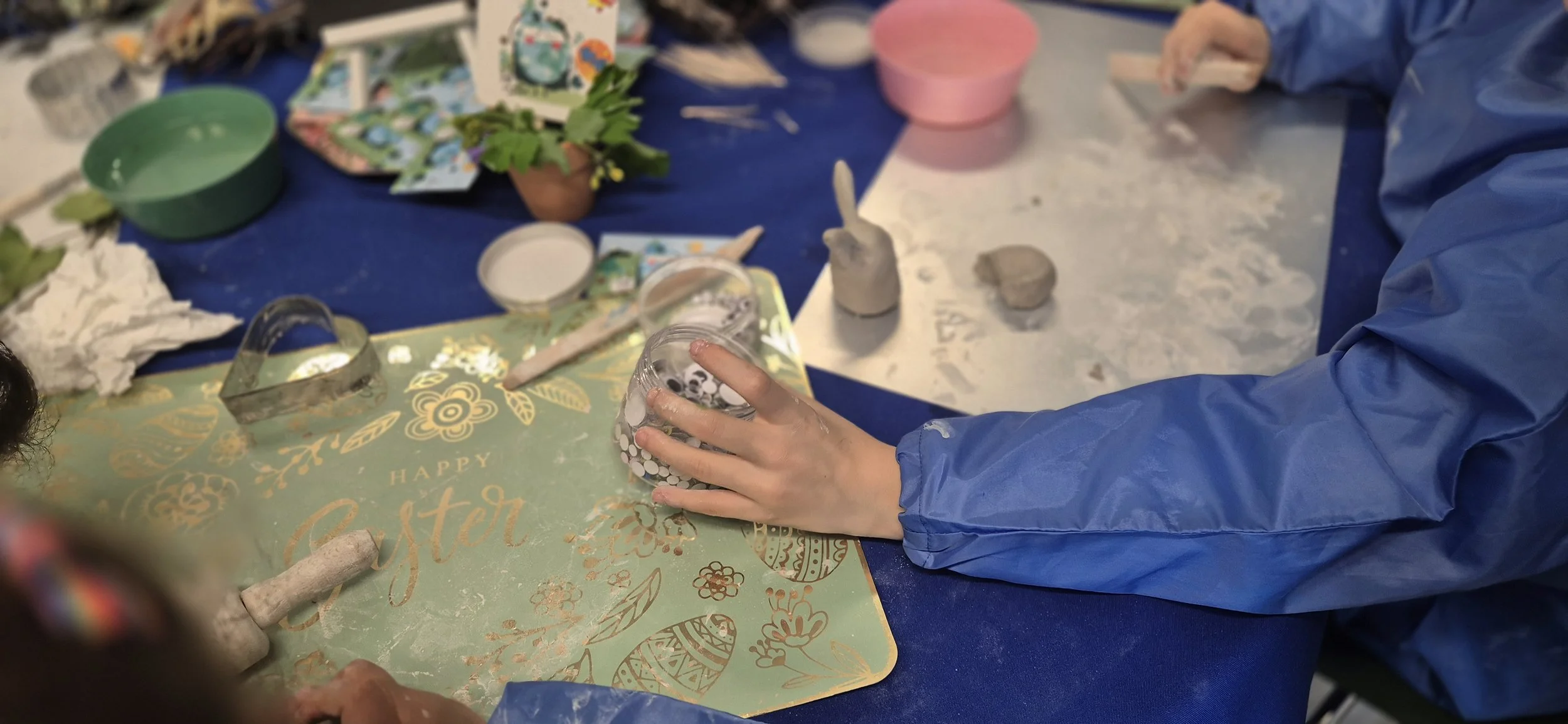 Une personne portant une veste bleue artisanale tenant un verre décoré avec des motifs blancs, entourée d'outils et de matériaux pour créer des objets en céramique sur une table couvre-plat bleu.