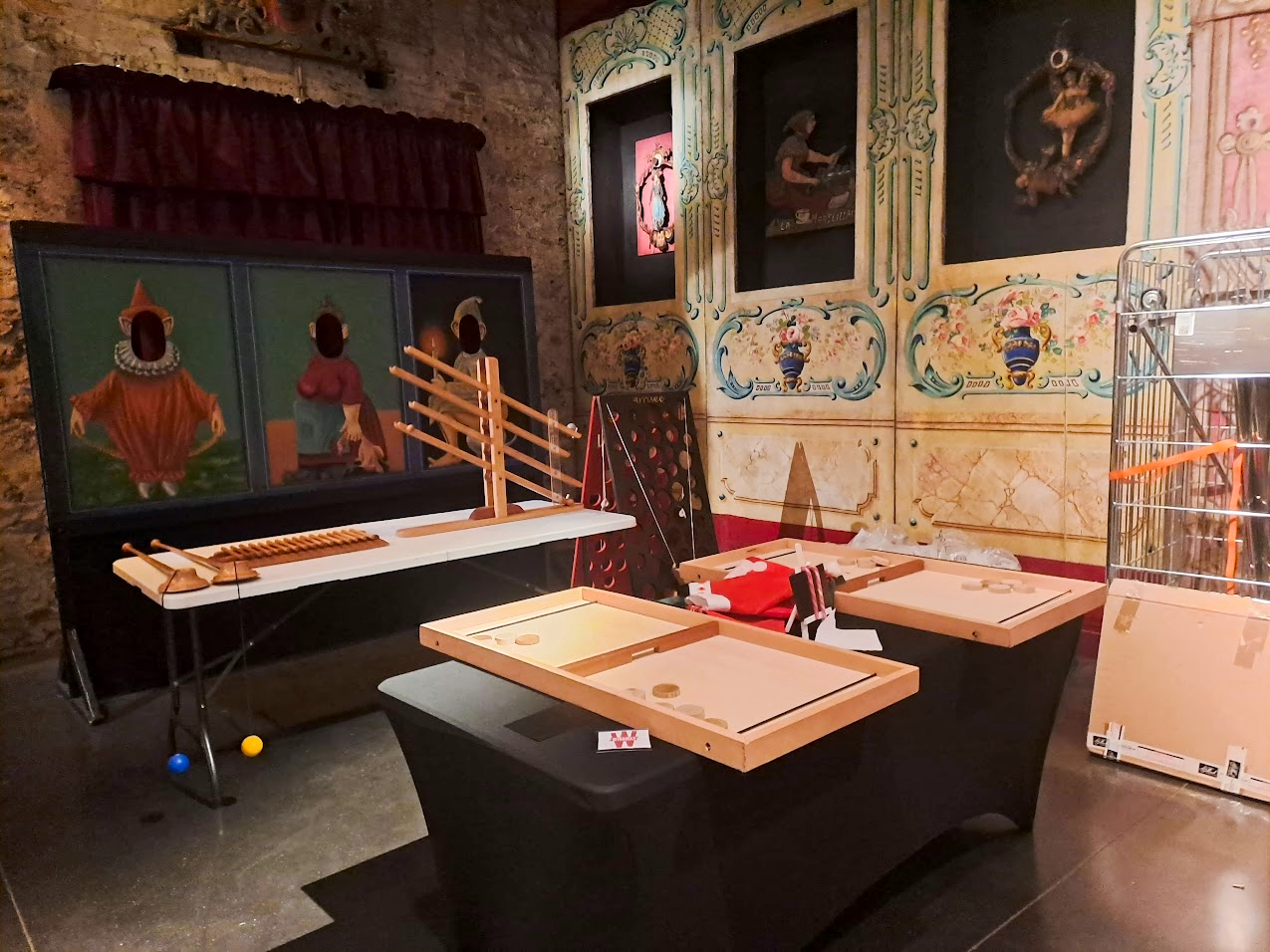 Une pièce avec des jeux de société et jouets anciens, décorée avec des fresques murales illustrant des scènes artistiques. On voit un tableau avec des personnages et des objets colorés, ainsi qu'un ensemble de jeux en bois, dont un jeu de palets et un mur d'échecs en bois.