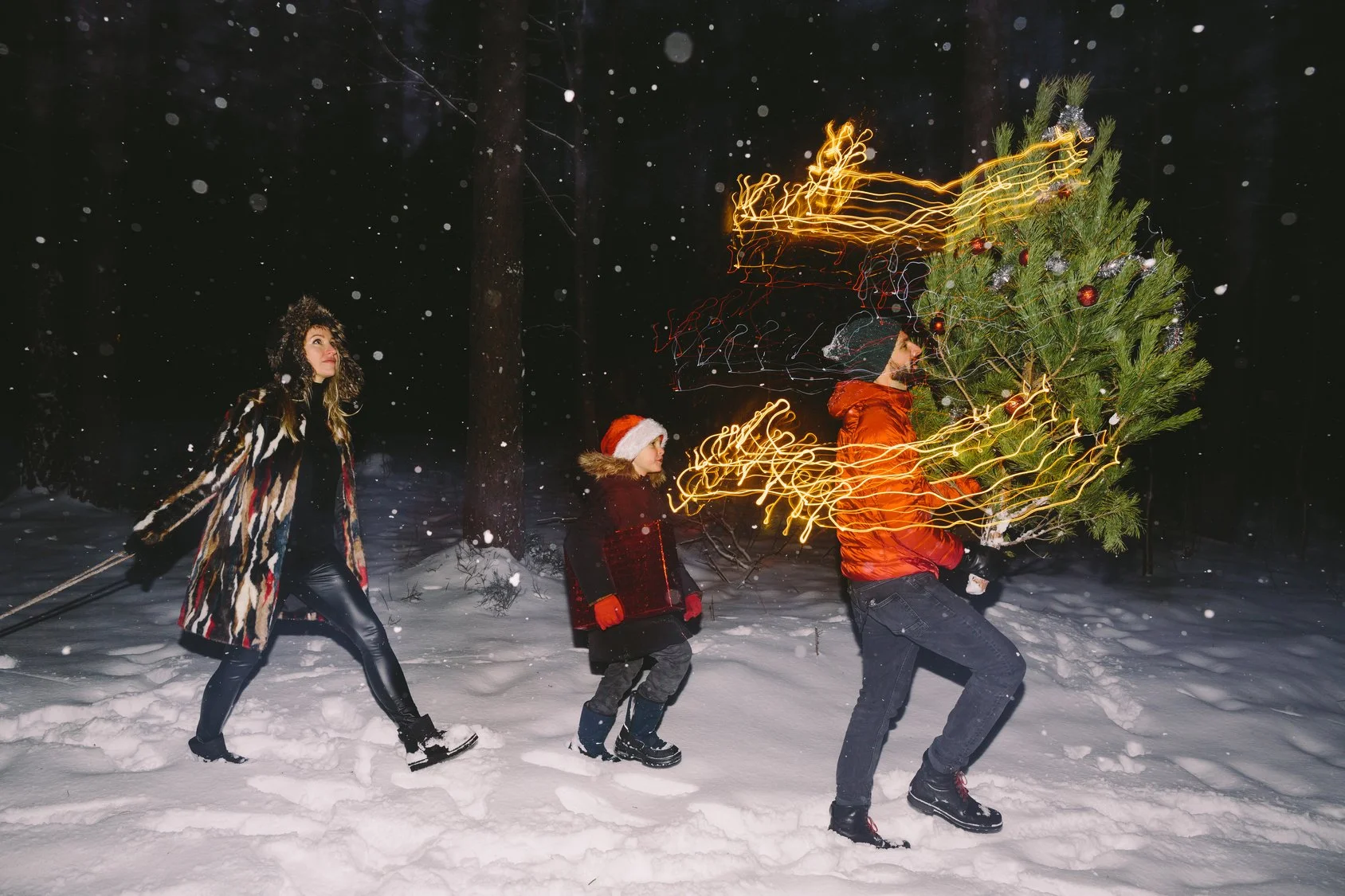 Une famille fait du tir à la corde entre deux personnes pendant qu'ils décorent un arbre de Noël dans la neige la nuit, avec des lumières orangées capturées en mouvement.