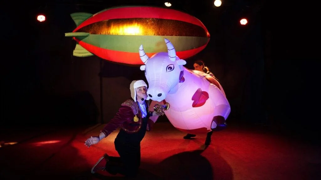 Crapaud géant gonflable avec une femme en costume assise sur son dos, scène de spectacle ou de théâtre avec éclairage dramatique.