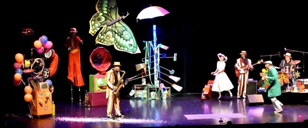 Une scène de spectacle avec des musiciens et des danseurs colorés sur scène, décoré de ballons, d'une grande papillon et d'objets ludiques.