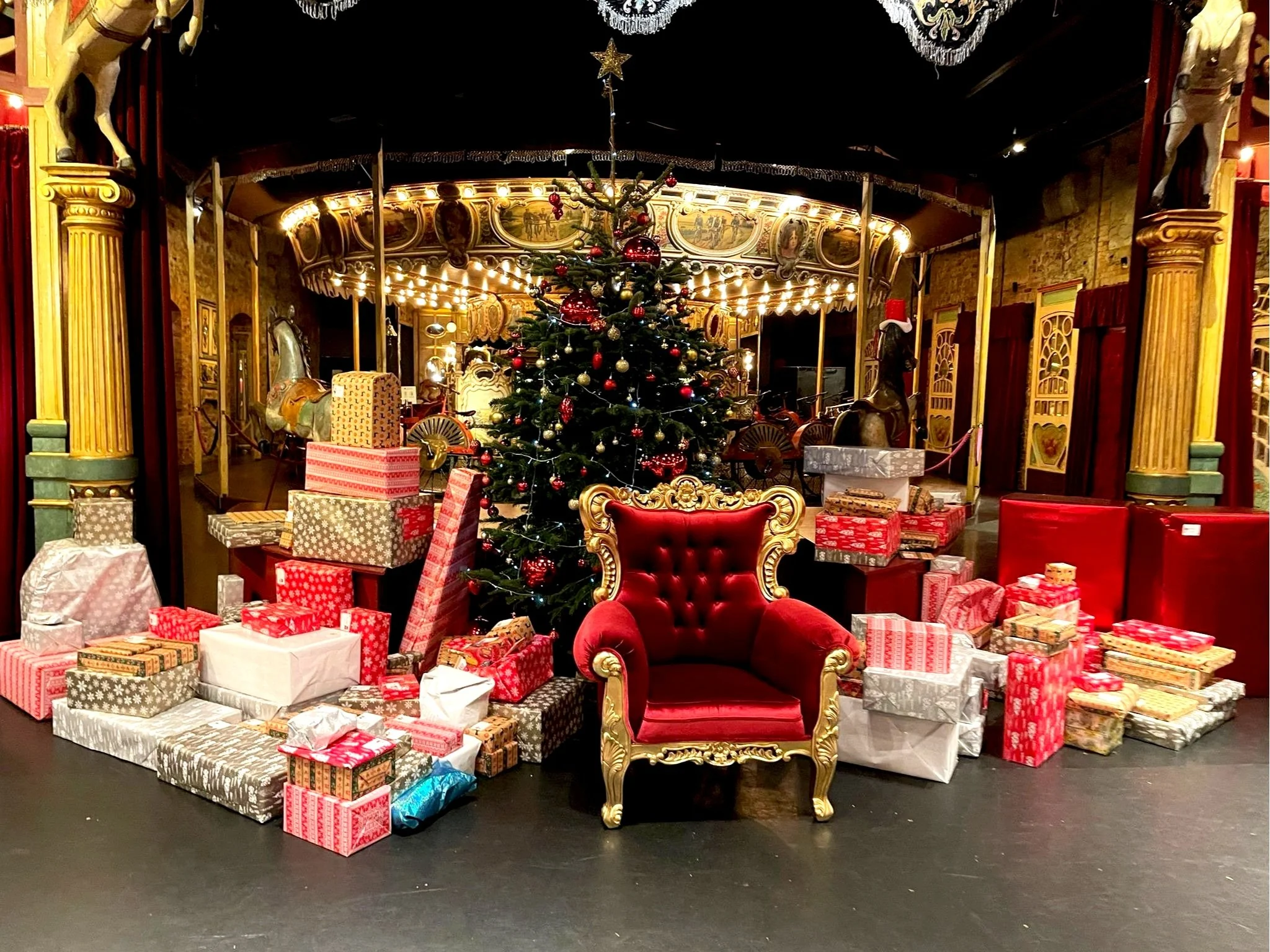 trone de Noël décoré entouré de cadeaux, avec un fauteuil rouge en velours à côté, dans une salle de spectacle ou un théâtre décoré pour Noël.