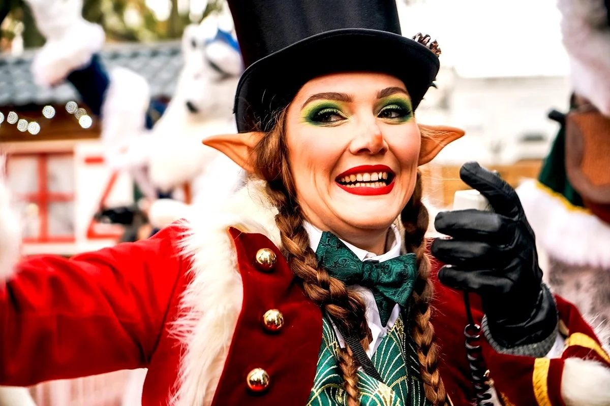 Femme déguisée en elfe avec oreilles pointues, vêtue d'un costume rouge festif avec fourrure et boutons dorés, portant un chapeau haut de forme noir, maquillage coloré, souriant en faisant un signe de la main, lors d'une fête ou parade