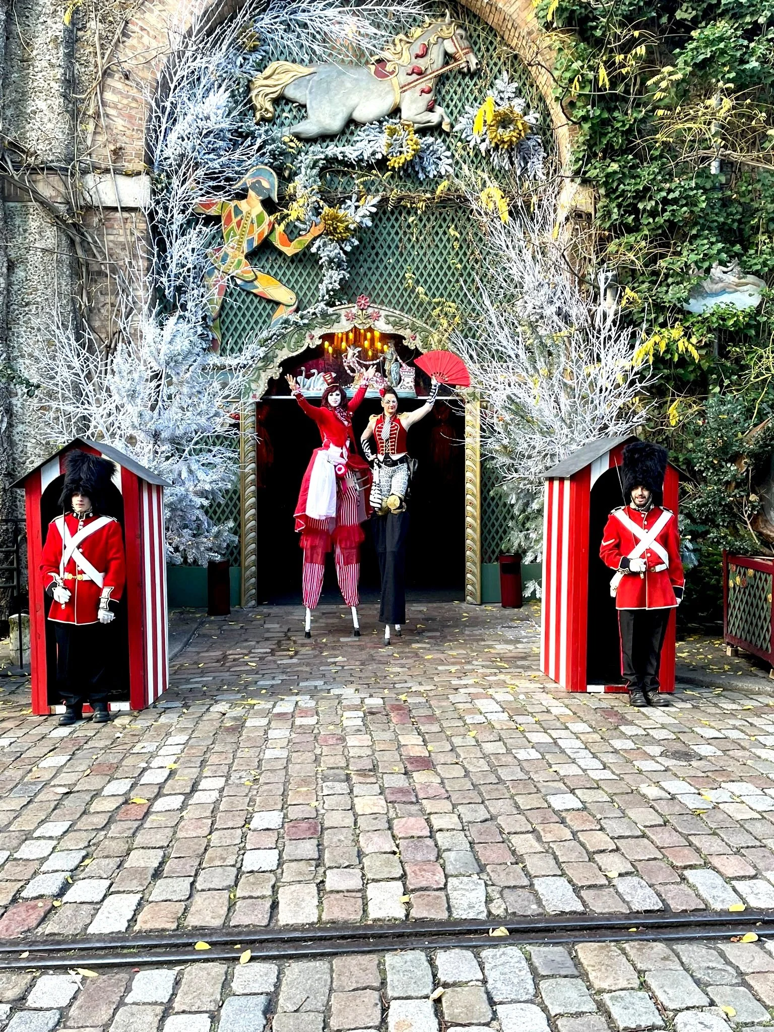 Décor de Noël avec deux soldats en tenue rouge et noirs, deux personnages en costumes de clown et de dame en costume rouge, arbres enneigés en décoration, chevaux en relief au-dessus, et fond rouge avec des lumières