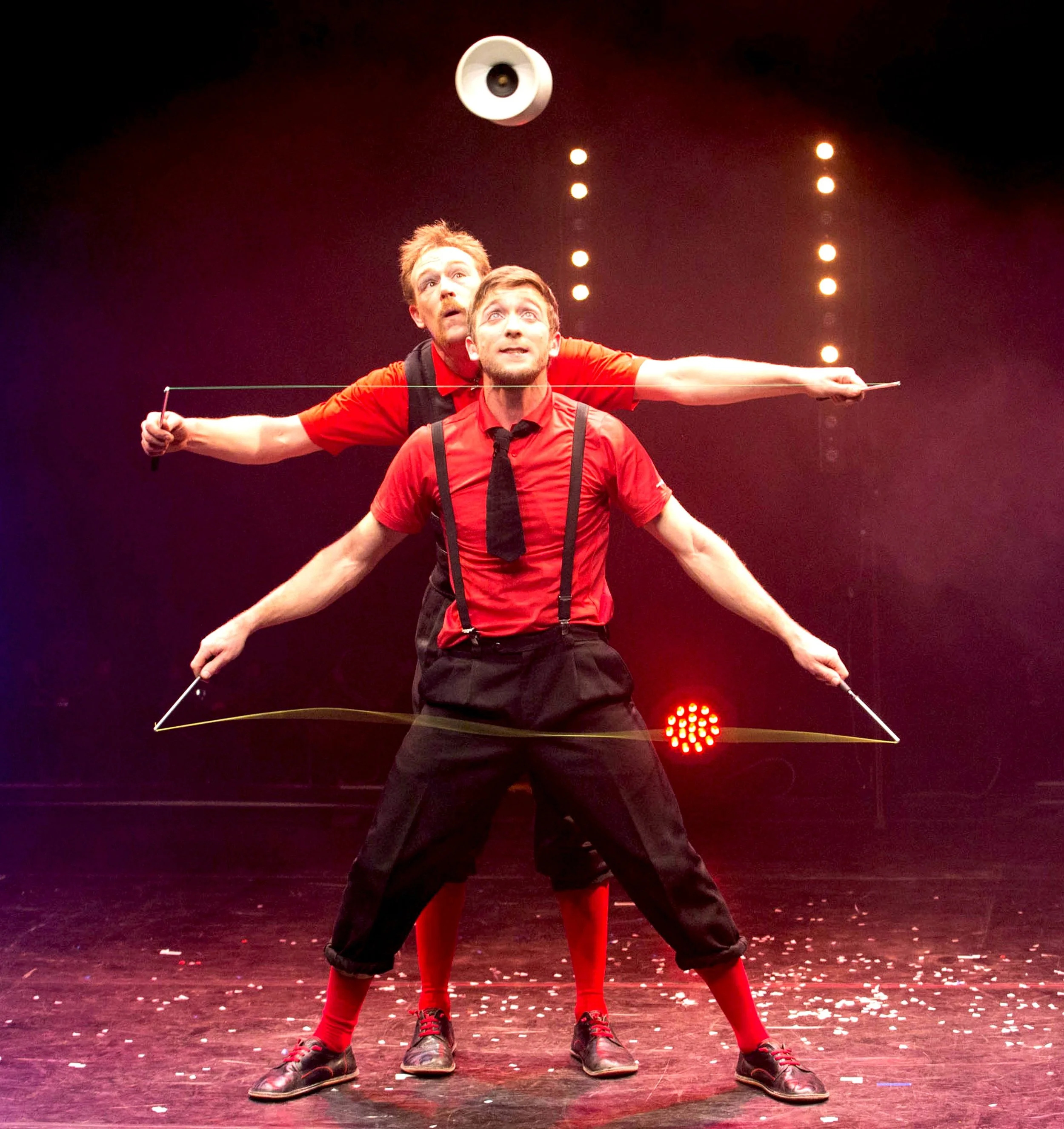 Deux hommes en costume noir et rouge exécutent un numéro de jonglerie avec des rubans dans un théâtre, avec des lumières de scène en arrière-plan.