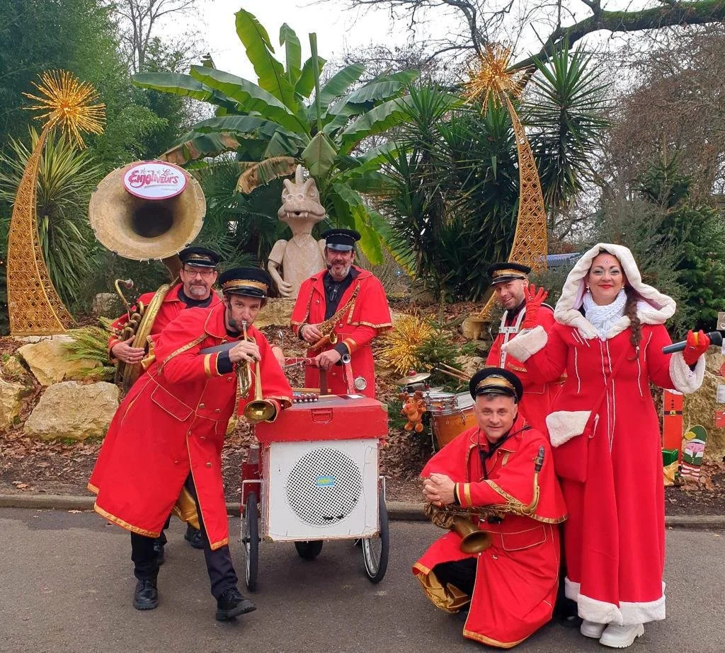 Groupe de musiciens déguisés en costumes rouges avec des accessoires de carnaval, jouant de divers instruments devant une décoration de carnaval avec des éléments festifs et végétation luxuriante.