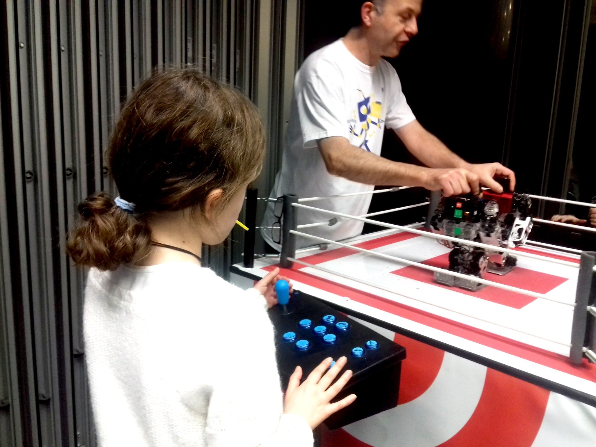 Une jeune fille joue à une console de jeu lors d'une démonstration de robotique tandis qu'un homme contrôle un robot sur un terrain de jeu.