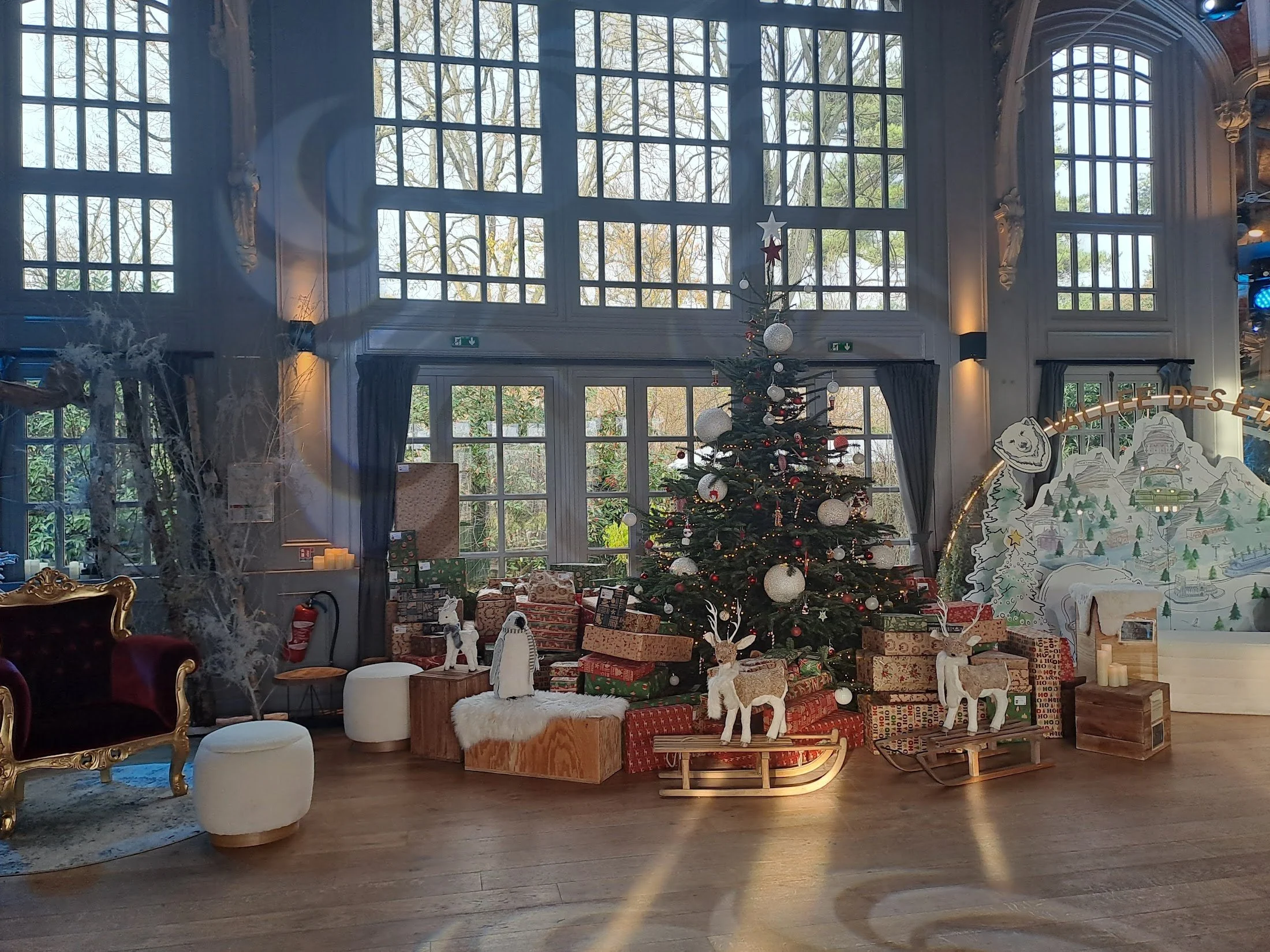 Décor de Noël avec un sapin décoré, cadeaux, renne en bois, et autres décorations dans une grande salle lumineuse.