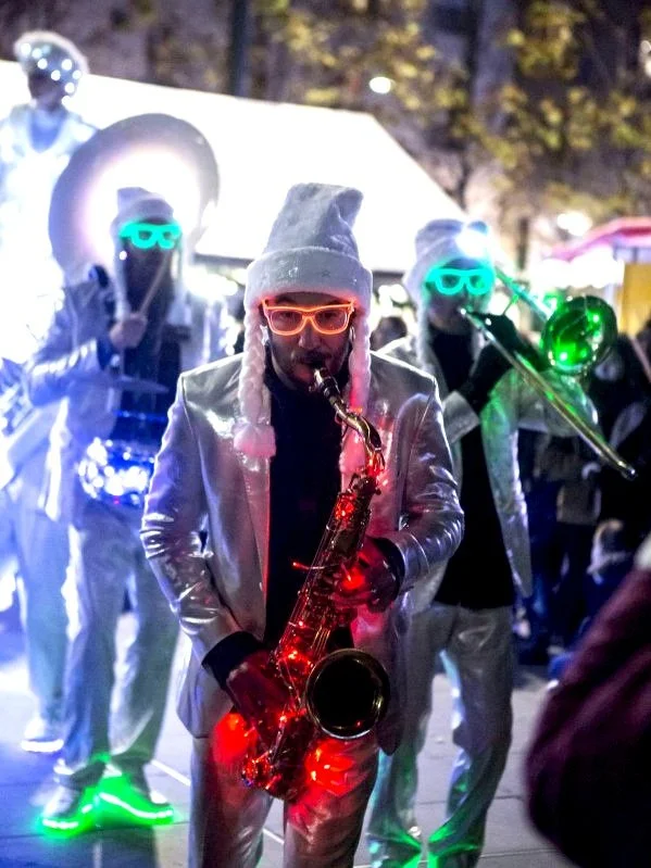 Groupe de musiciens en costume brillant avec des lunettes lumineuses et des instruments de musique, jouant lors d'une performance nocturne en extérieur.