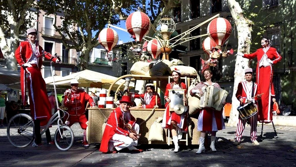 Un groupe de musiciens et performers en costumes rouges et blancs, avec des lampions rouges et blancs suspendus, dans une place urbaine avec des arbres et des bâtiments en arrière-plan.