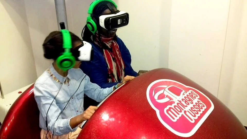 Deux jeunes filles portant un casque de réalité virtuelle et un casque audio vert, assises devant une table rouge avec un logo 'Montagnes Russes'.