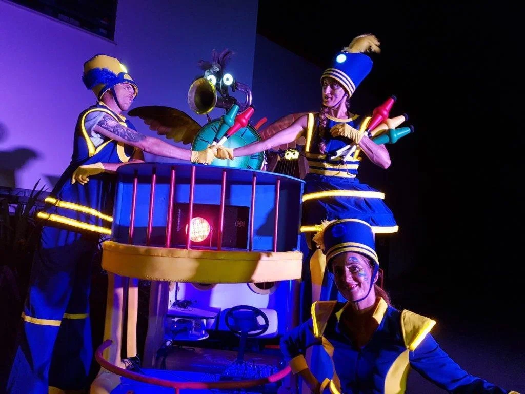 Des artistes déguisés en pompiers jouant avec des accessoires colorés lors d'une performance artistique ou d'un spectacle. L'ambiance est colorée et lumineuse.