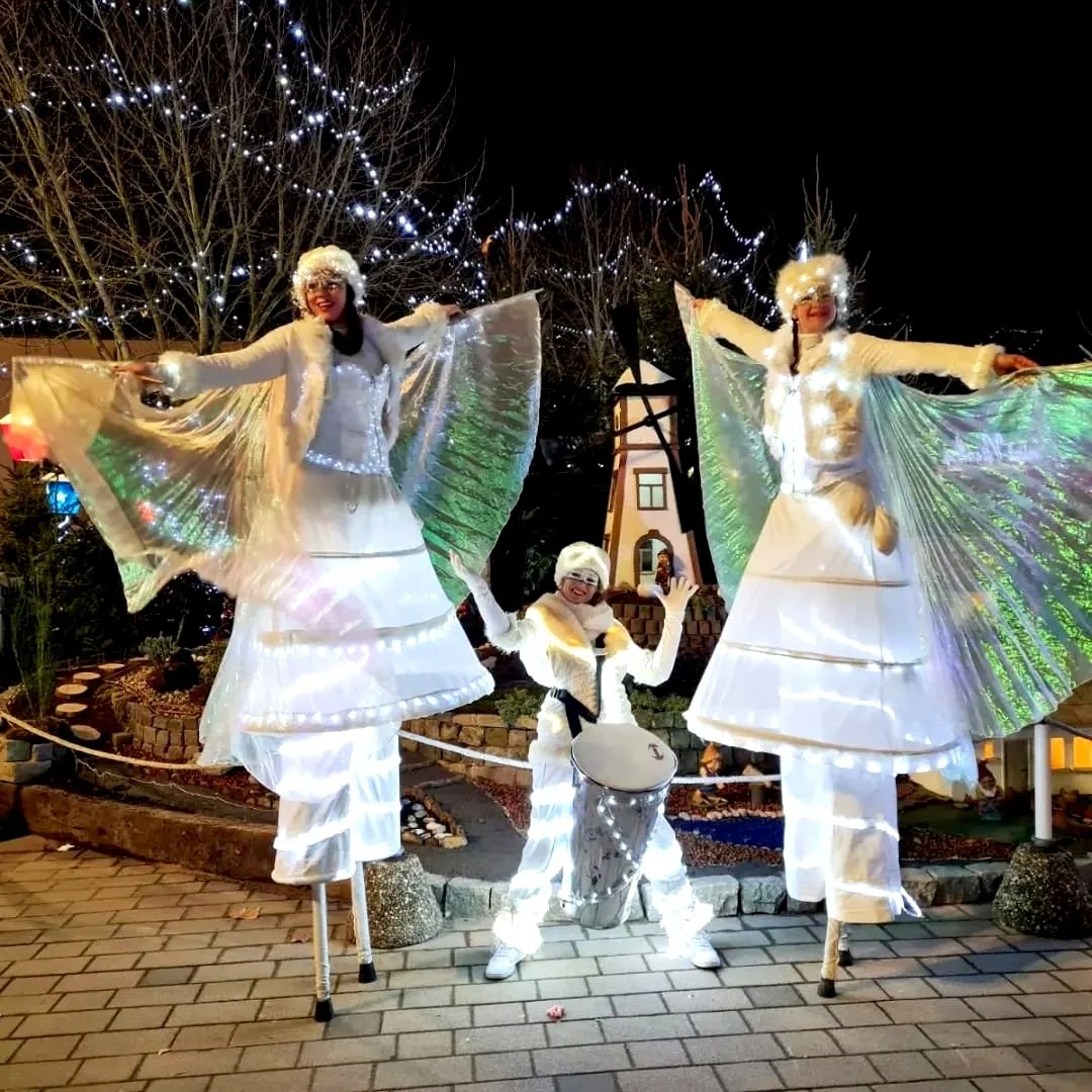 Trois personnes déguisées en anges sur des échasses, portant des costumes blancs avec des ailes lumineuses, lors d'une fête nocturne avec des décorations de lumières en arrière-plan.