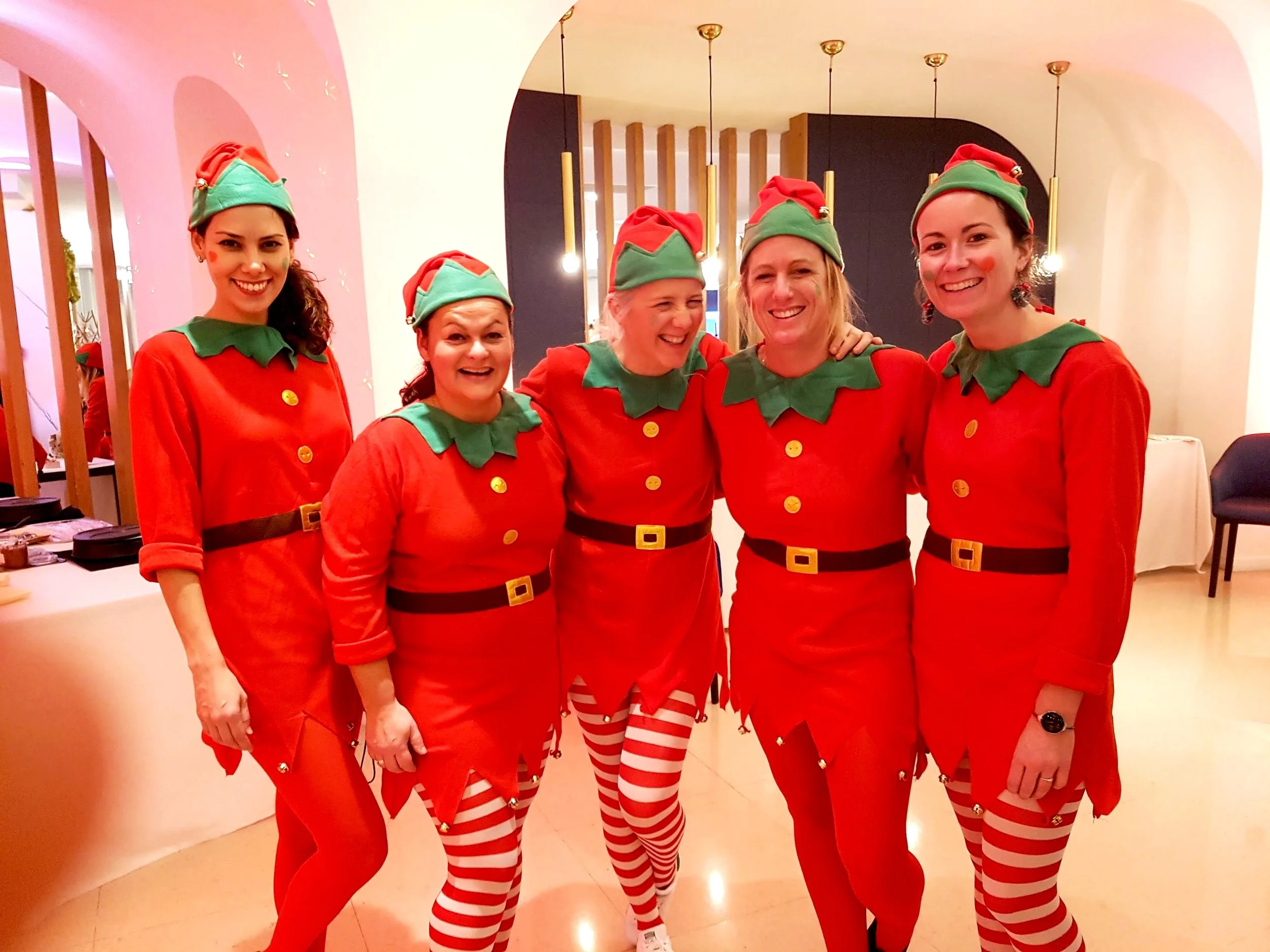 Groupe de cinq femmes déguisées en lutins de Noël, portant des costumes rouges avec des collants à rayures rouges et blanches, des chapeaux verts et rouges et des costumes assortis avec des ceintures noires et des colliers verts.