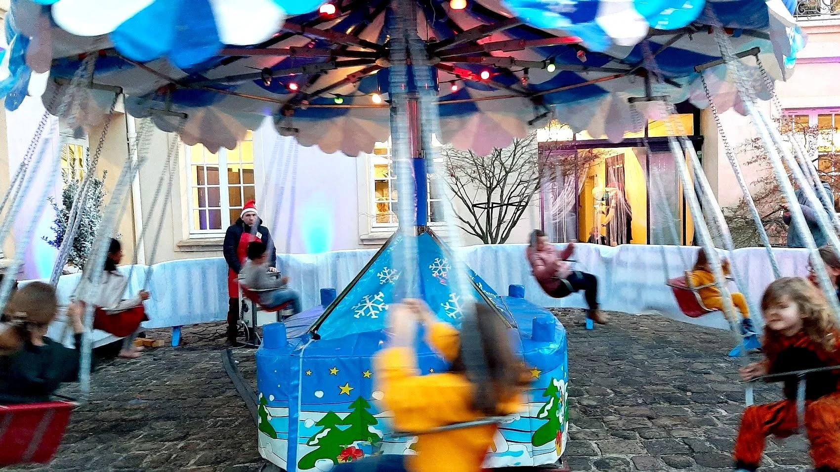 Une fête foraine avec une grande roue en rotation, décorée de motifs de flocons de neige, de sapins et de motifs festifs, avec des enfants qui tournent sur des sièges suspendus, dans un environnement extérieur devant une maison.