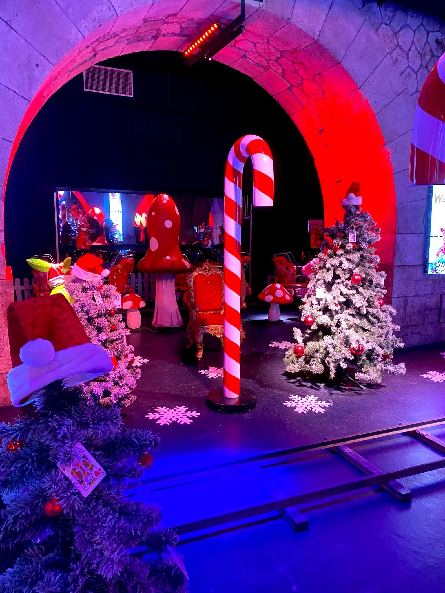 Décor de Noël avec deux sapins décorés, un champignon géant, un bonbon géant, un fauteuil rouge, et un escalier de queuille de gourmandise, sous un éclairage coloré rouge et bleu.