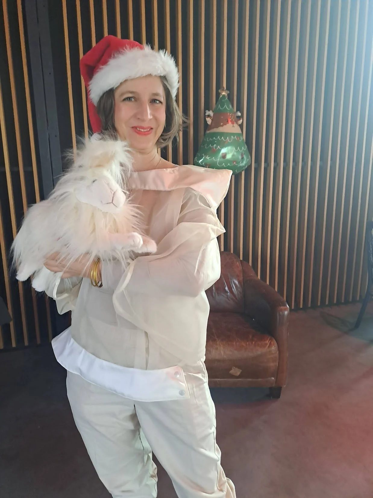 Femme portant un bonnet de Père Noël, tenant un chat blanc dans un décor de Noël avec un sapin en fond.