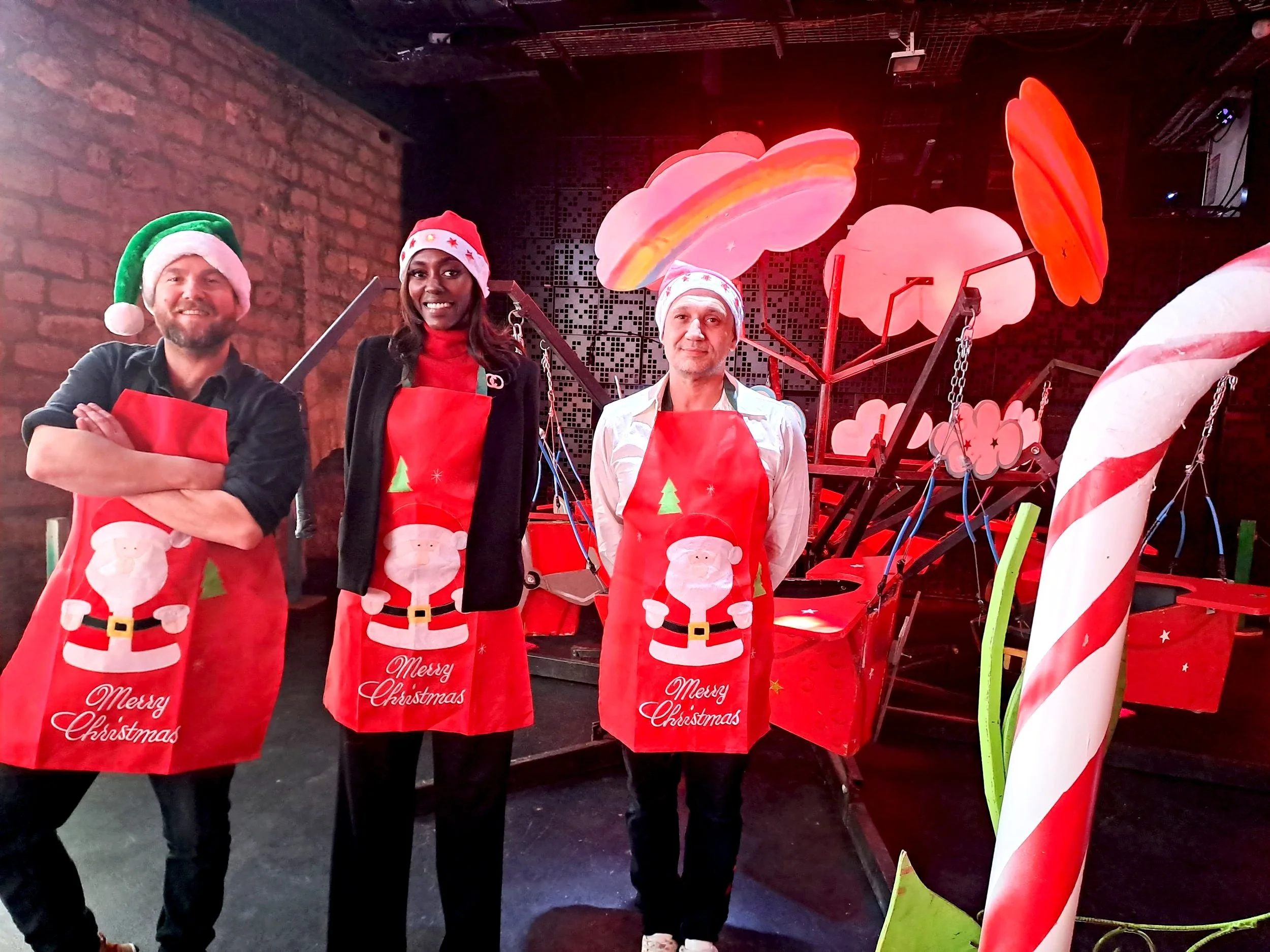 Trois personnes souriantes portant des tabliers de Noël avec un décor festif en arrière-plan. 