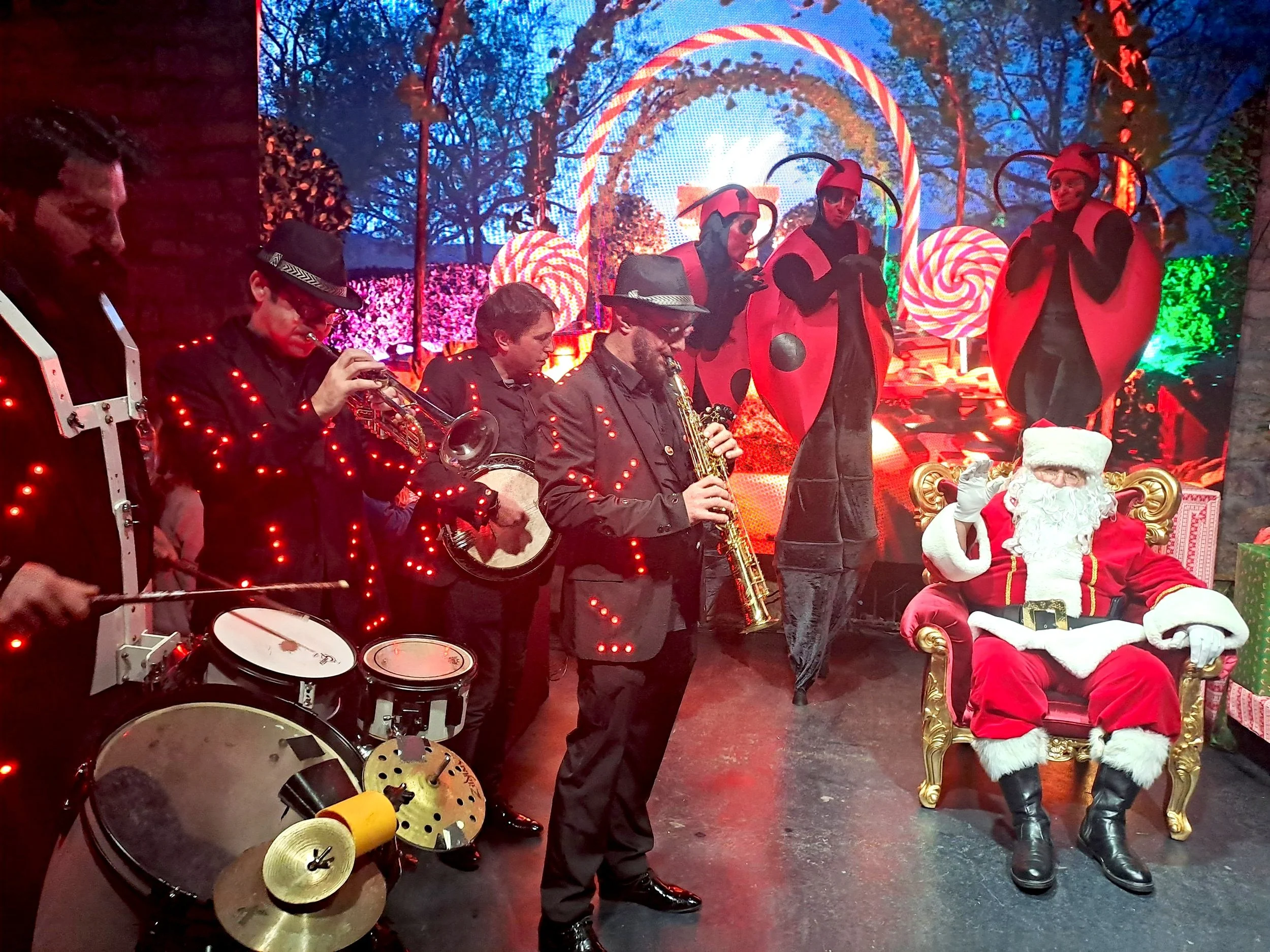 Un groupe de musiciens avec des instruments joue devant un décor de fête de Noël. Santa Claus est assis sur un fauteuil doré, portant un costume rouge avec une barbe blanche. En arrière-plan, des personnages en costumes de coccinelle avec de grands yeux et antennes.