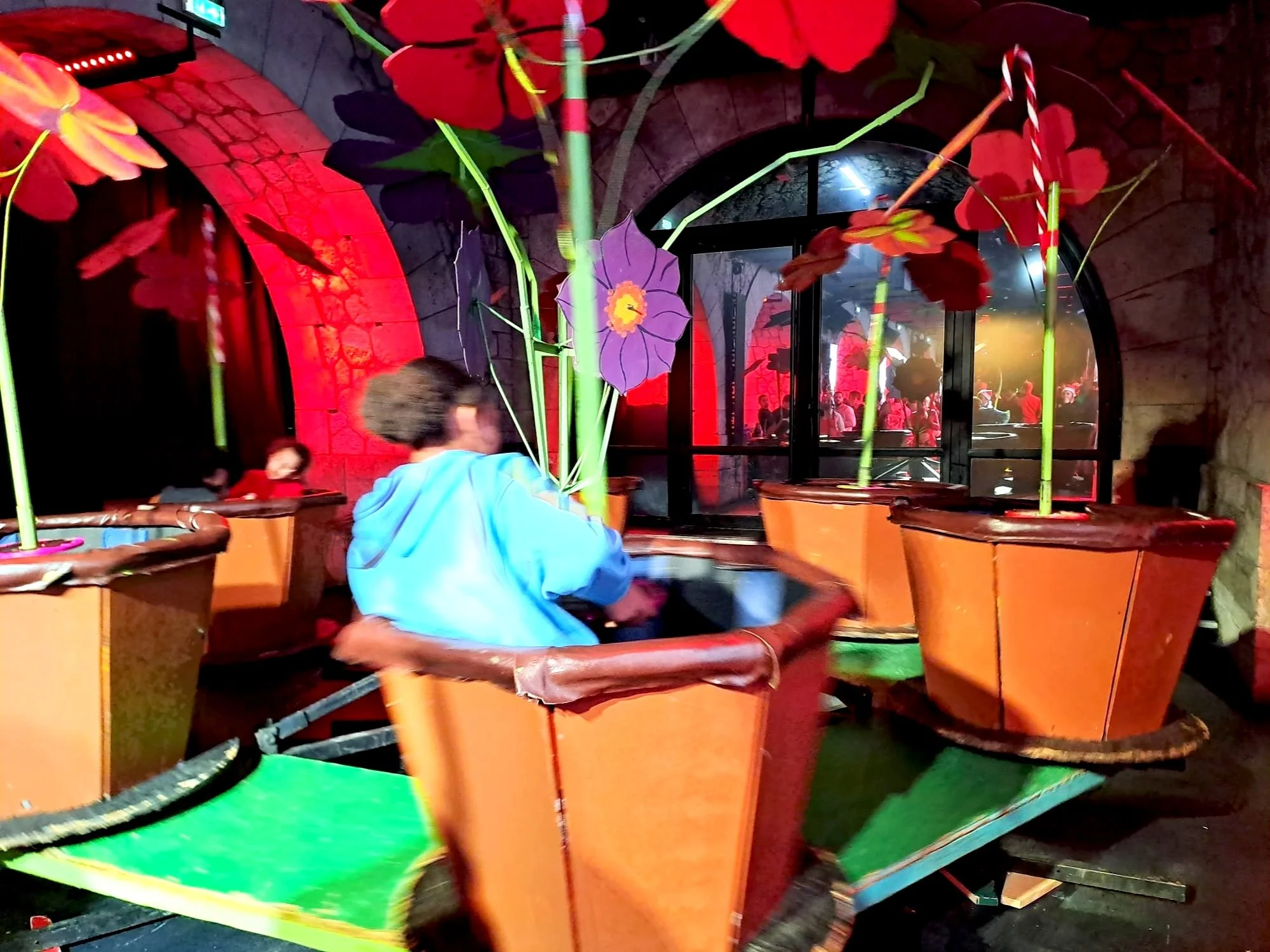 Une salle de fête ou danse avec des décorations colorées, notamment des fleurs en plastique dans des pots orange et des lumières vives. On peut voir des personnes en arrière-plan dans une atmosphère festive.