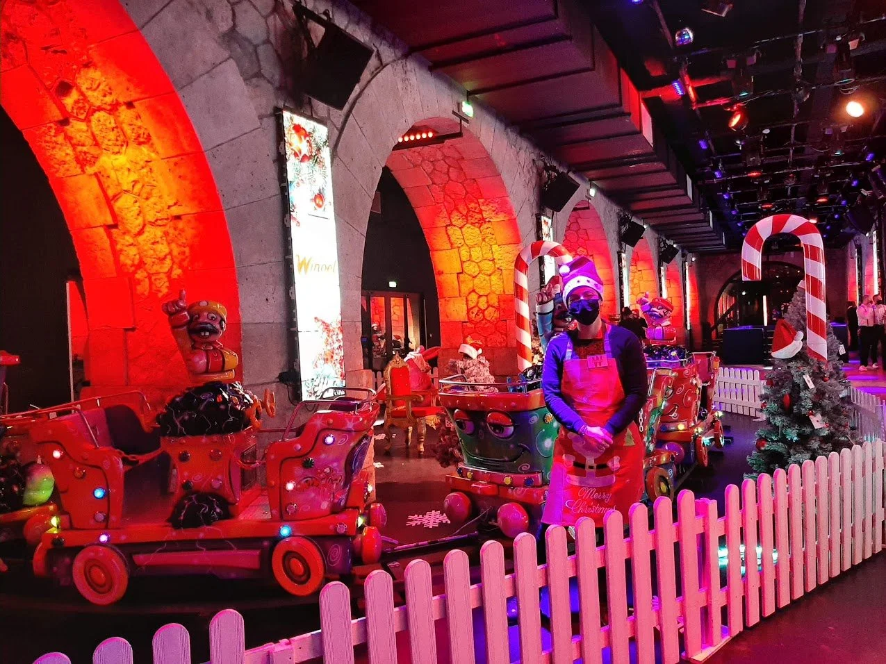 Décoration de Noël à l'intérieur avec des trains en bois, un sapin de Noël et des personnes portant des masques.