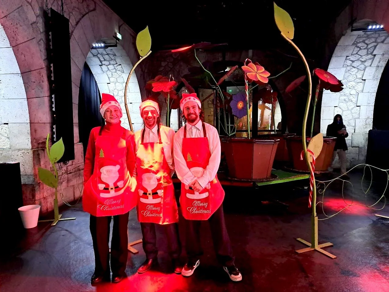 Trois personnes déguisées en Père Noël avec des tabliers de Noël, dans un décor festif de Noël avec des décorations de Noël et un traîneau derrière eux, dans une salle aux murs de pierre.