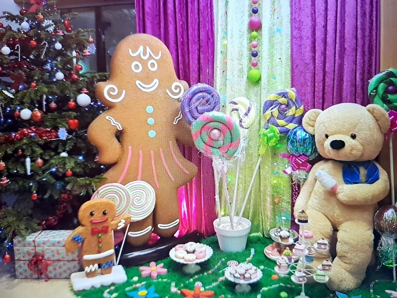 Décoration de Noël avec un sapin décoré d'ornements colorés, un grand biscuit en pain d'épice avec un visage souriant, un ours en peluche, des sucres d'orge, des bonbons, cupcakes et autres friandises colorées sur une table, avec des rideaux roses et vert clair en arrière-plan.