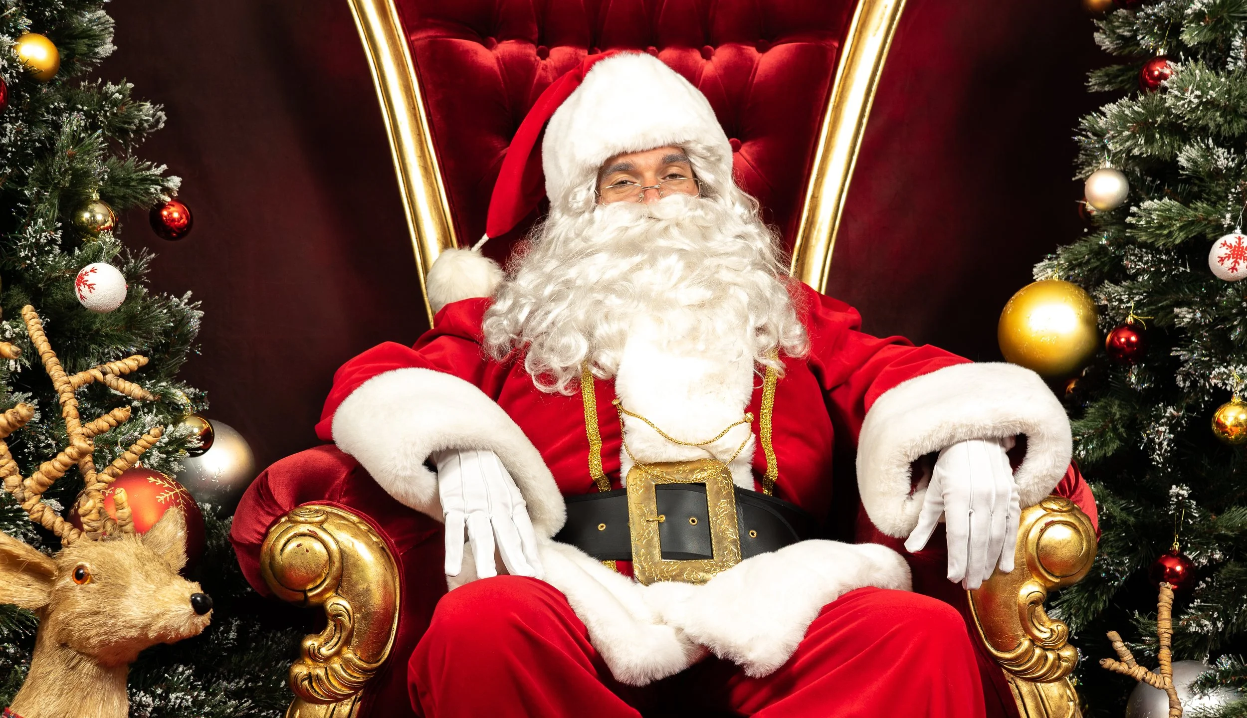 Papa Noël assis sur un fauteuil rouge, entouré de sapins décorés avec des boules de Noël, à côté d'une girafe en peluche, dans une scène festive de Noël.