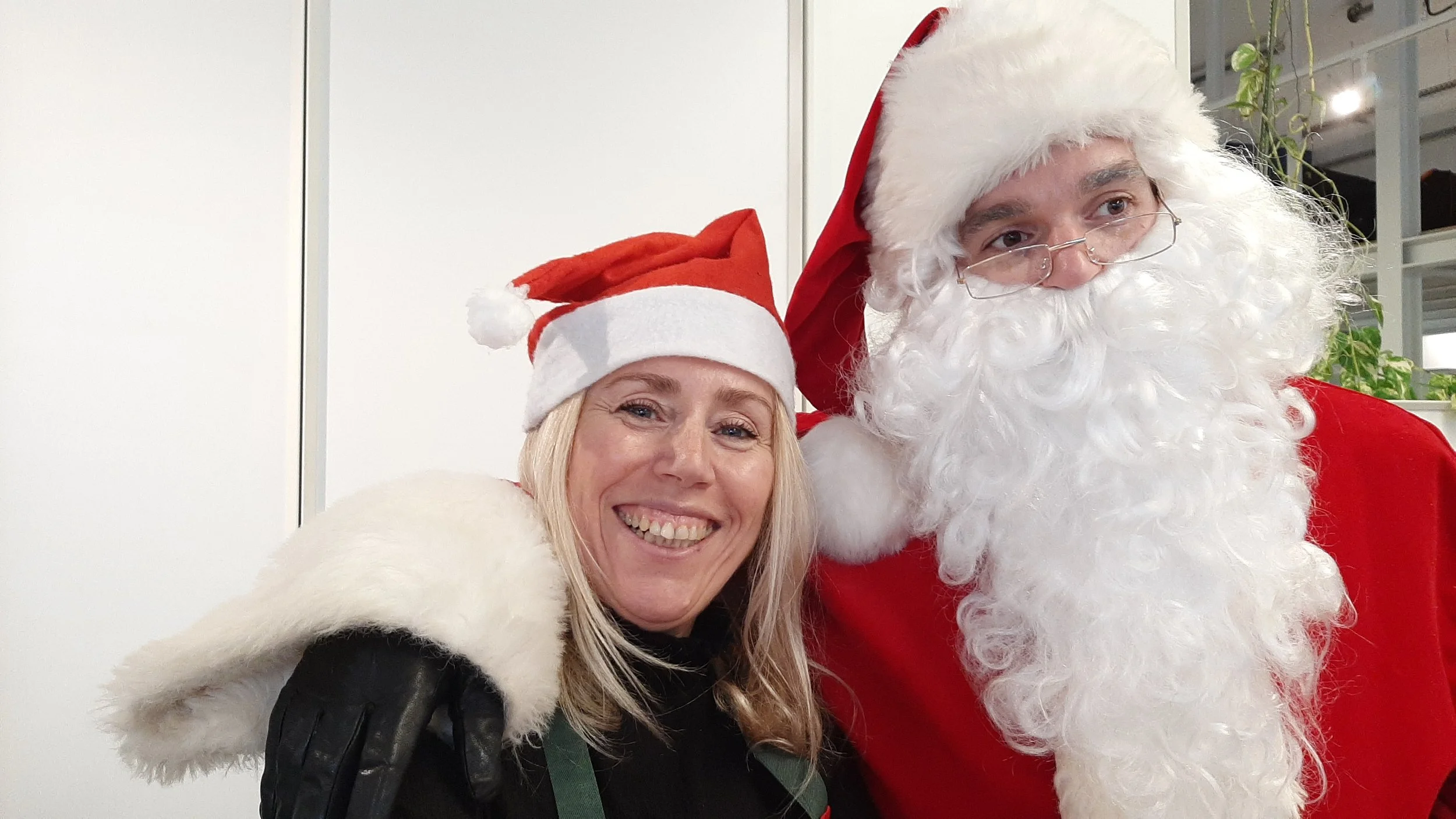 Une femme souriante portant un bonnet de Noël rouge et blanc, à côté du Père Noël, habillé de rouge avec une longue barbe blanche, dans un décor intérieur.