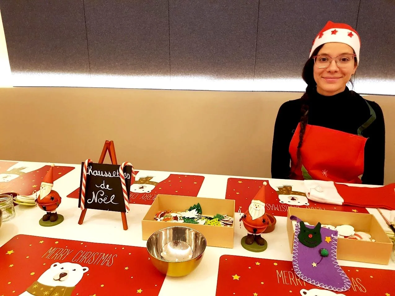 Une femme portant un bonnet de Noël rouge avec des étoiles rouges, debout derrière une table décorée pour Noël, avec des figurines et des décorations de Noël.