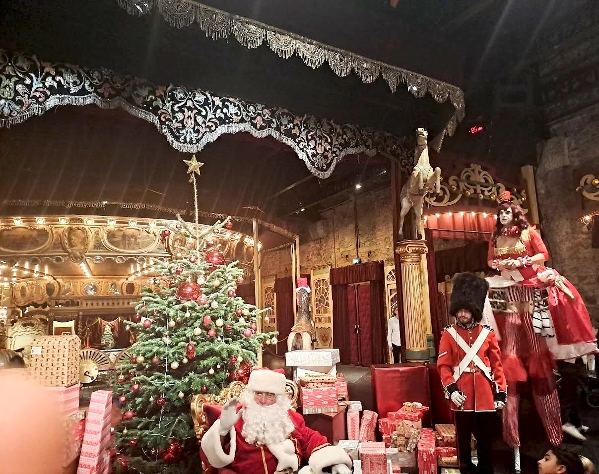Décor de fête de Noël avec un sapin décoré, Père Noël assis, soldats en costume royal et une jeune femme en costume de carnaval dans une salle ornée de décorations festives.