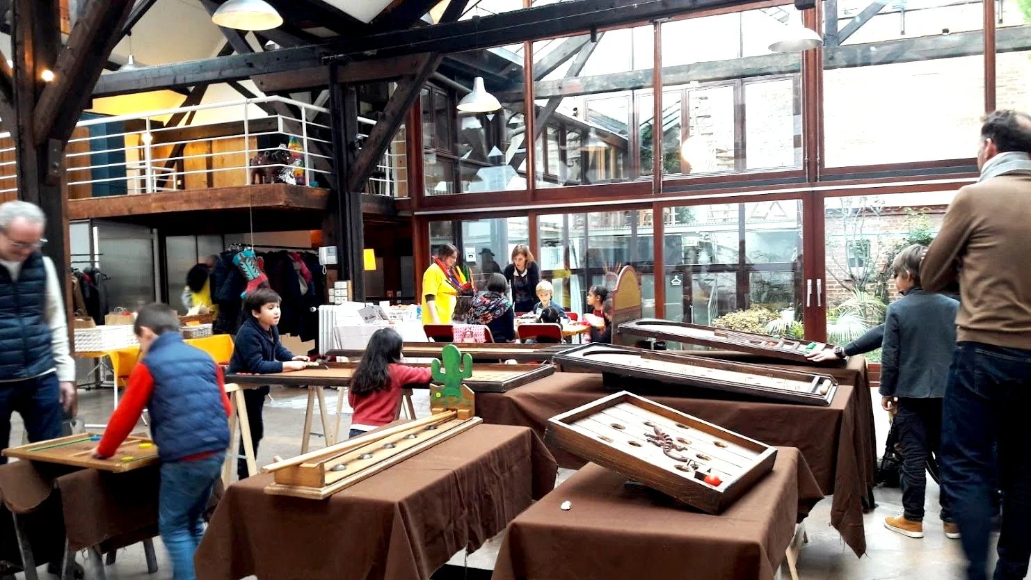 Une salle avec plusieurs tables couvertes de tissus marron, où des enfants et des adultes jouent à des jeux de société et de société. La pièce a de grandes fenêtres en verre laissant entrer beaucoup de lumière naturelle. Au fond, il y a une vitrine avec des vêtements et un comptoir avec des personnes achetant ou réglant des affaires.