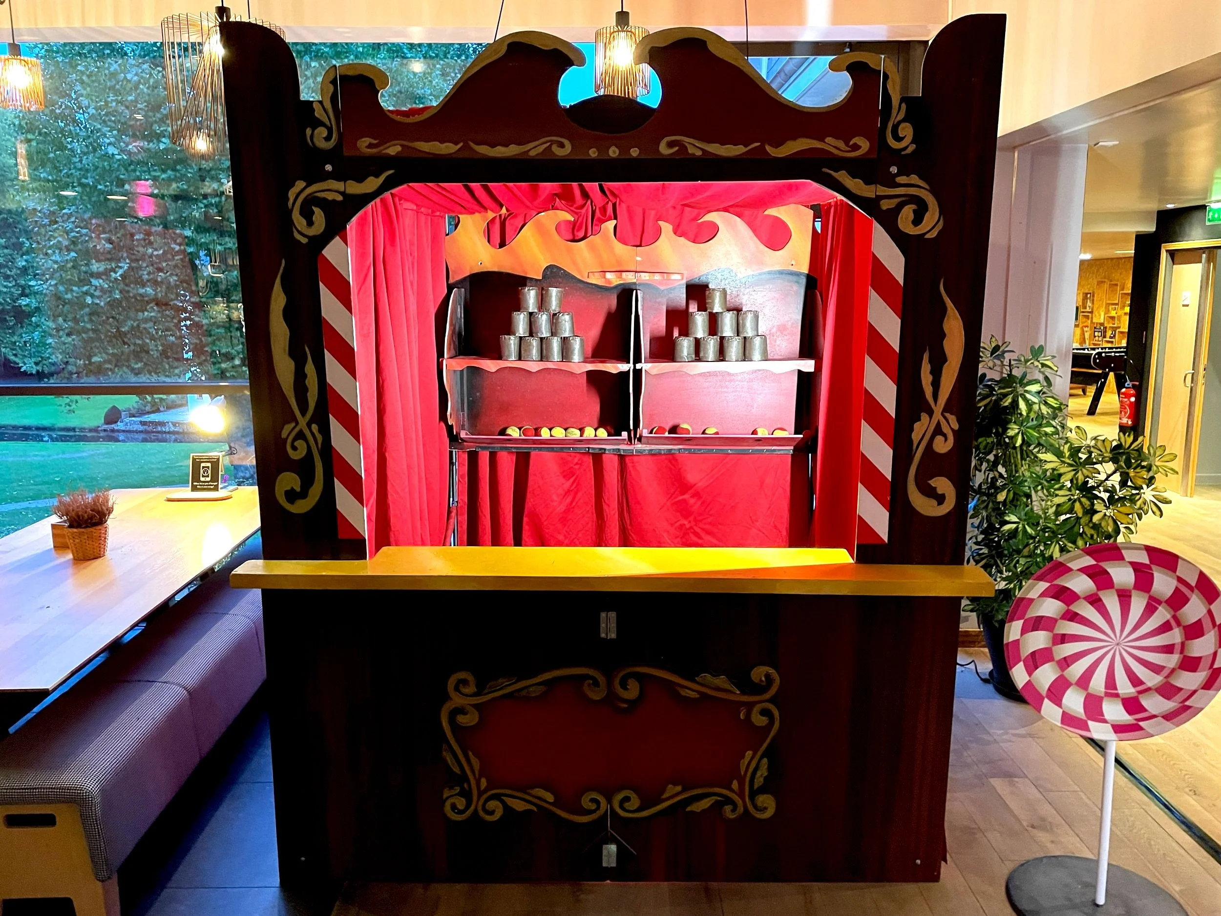 Un théâtre de marionnettes en bois avec une scène rouge, décorée de motifs dorés, contenant des boîtes métalliques empilées et des balles colorées. Dehors, à gauche, il y a une table en bois avec une petite plante en pot; à droite, un stand de bonbons en forme de cercle avec un motif rose et blanc. Le fond montre une fenêtre avec des arbres et une lumière extérieure.
