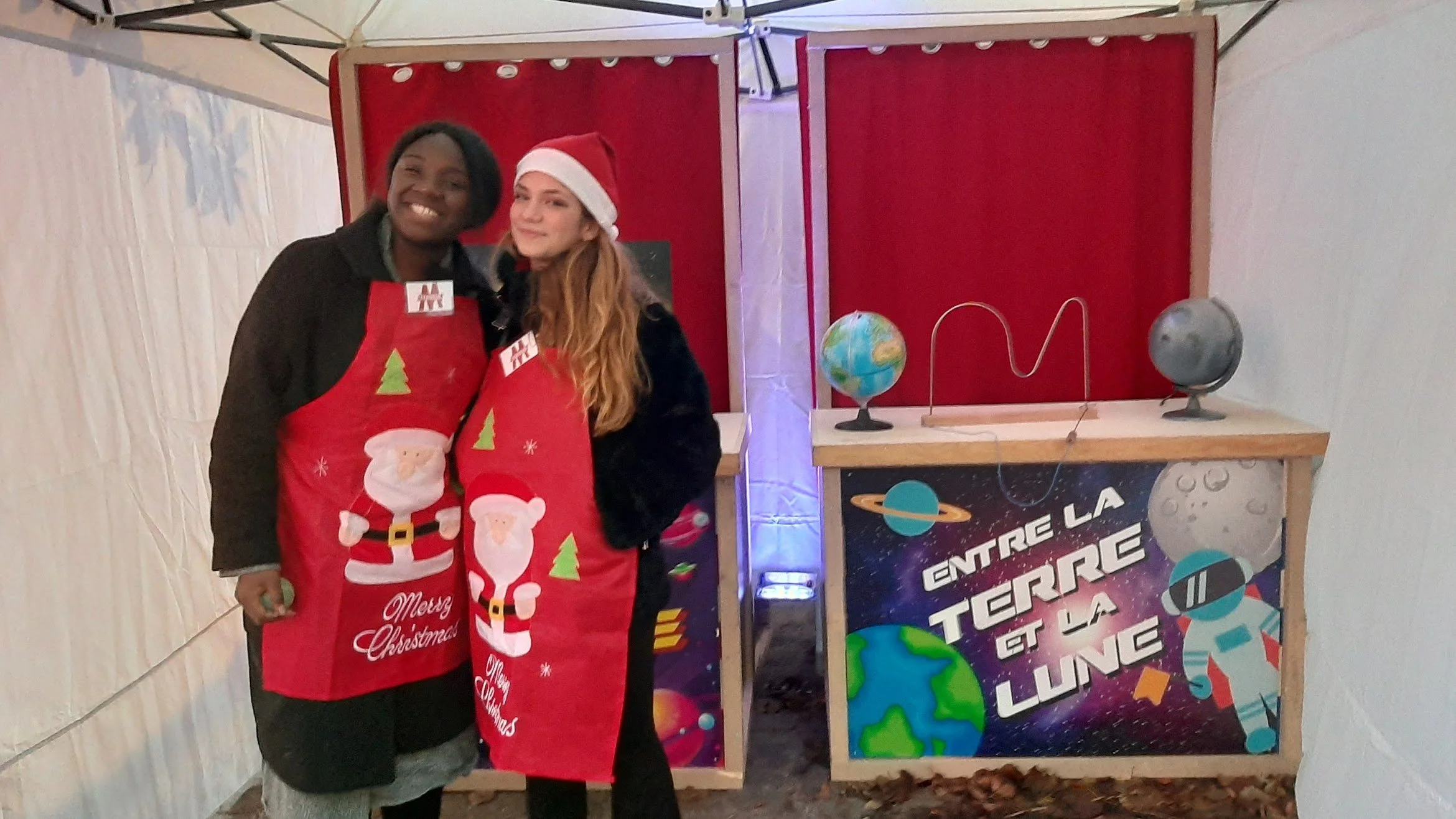 Deux jeunes femmes portant des tabliers de Noël rouges avec des motifs de Père Noël et de sapin, posant devant une affiche sur le thème de l'espace avec la phrase "Entre la Terre et la Lune".