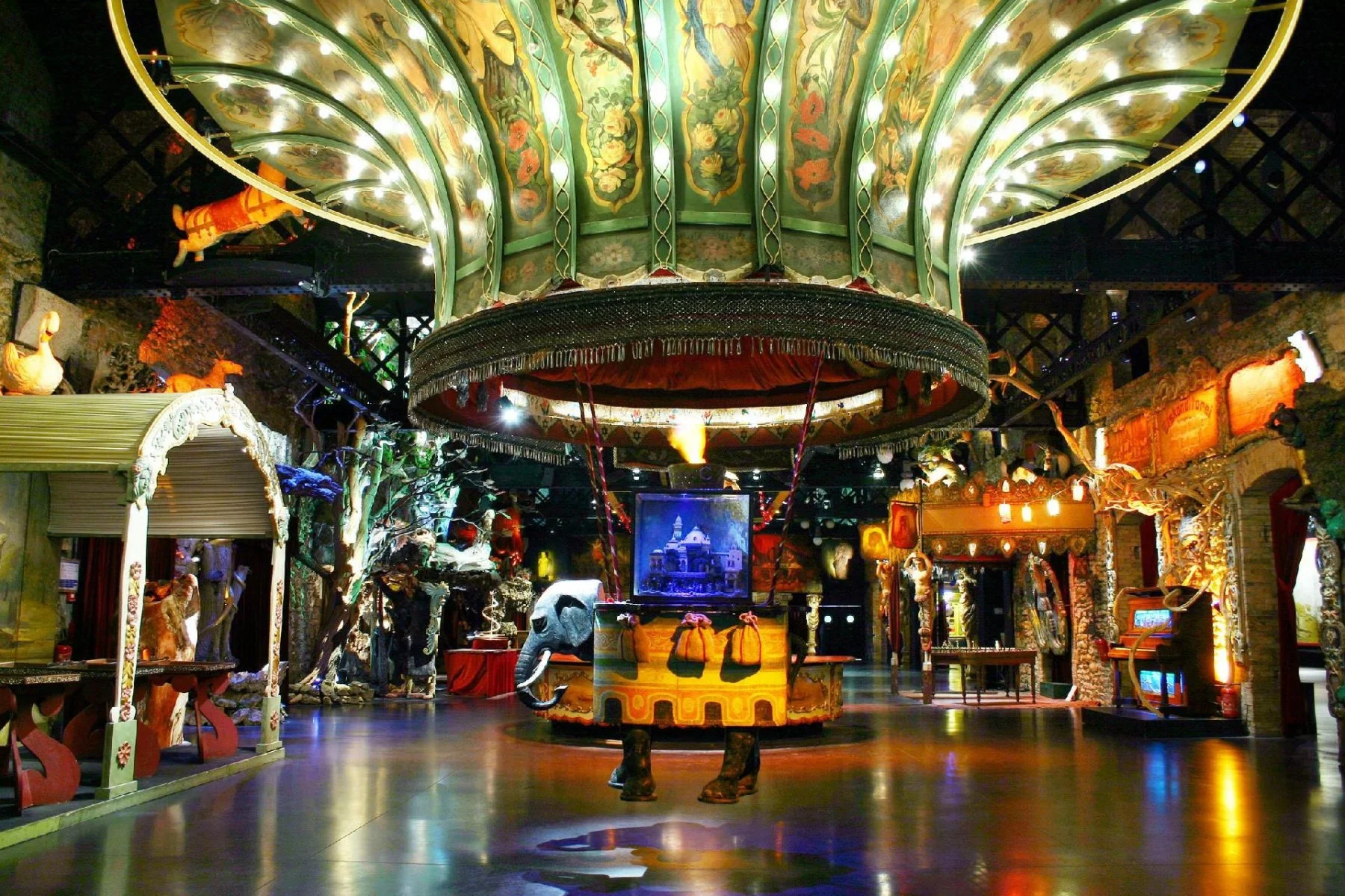 Une salle intérieure décorée avec un manège sculpté, des statues d'animaux en peluche, et diverses décorations colorées, évoquant un style féerique ou de carnaval.